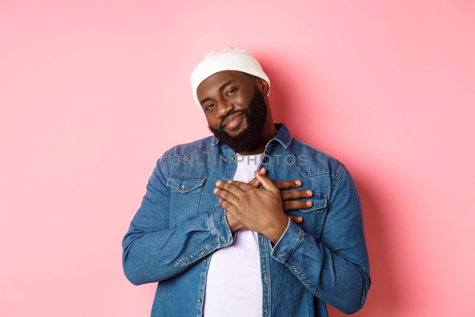 Thankful and touched Black guy saying thank you, smiling adorable and holding hands on heart, appreciate nice gesture, standing over pink background by Benzoix