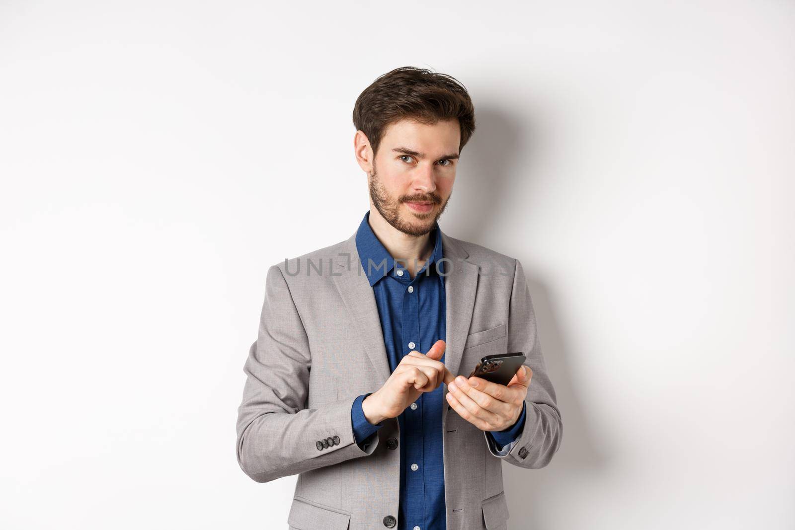 Handsome bearded male model in suit using mobile phone, smiling pleased at camera, white background by Benzoix