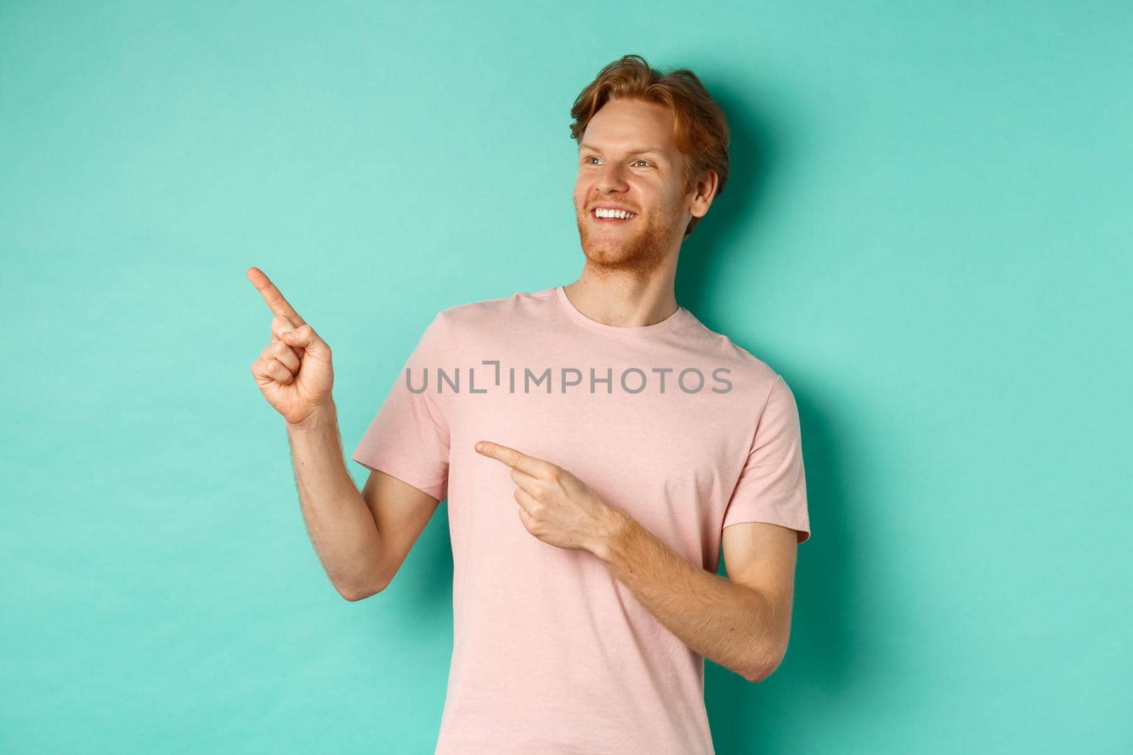 Handsome smiling man with red hair and beard looking delighted, pointing at upper left corner banner, standing in t-shirt over mint background.