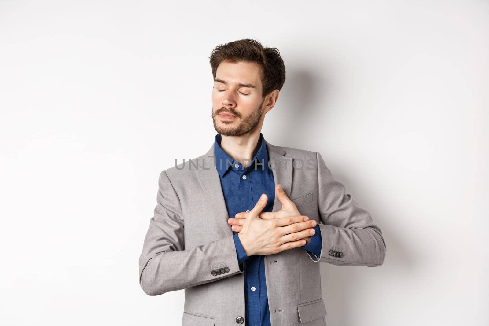 Romantic bearded man in stylish suit holding hands on heart, close eyes and remember something with tender feelings, standing nostalgic on white background.