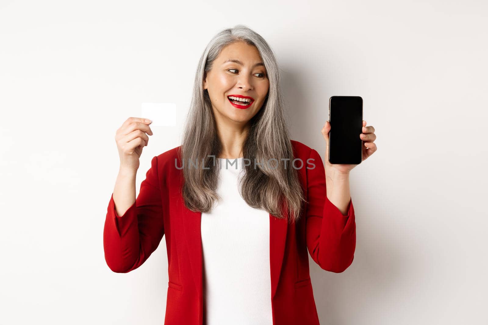 Cheerful asian mature woman showing blank smartphone screen and credit card, concept of e-commerce.