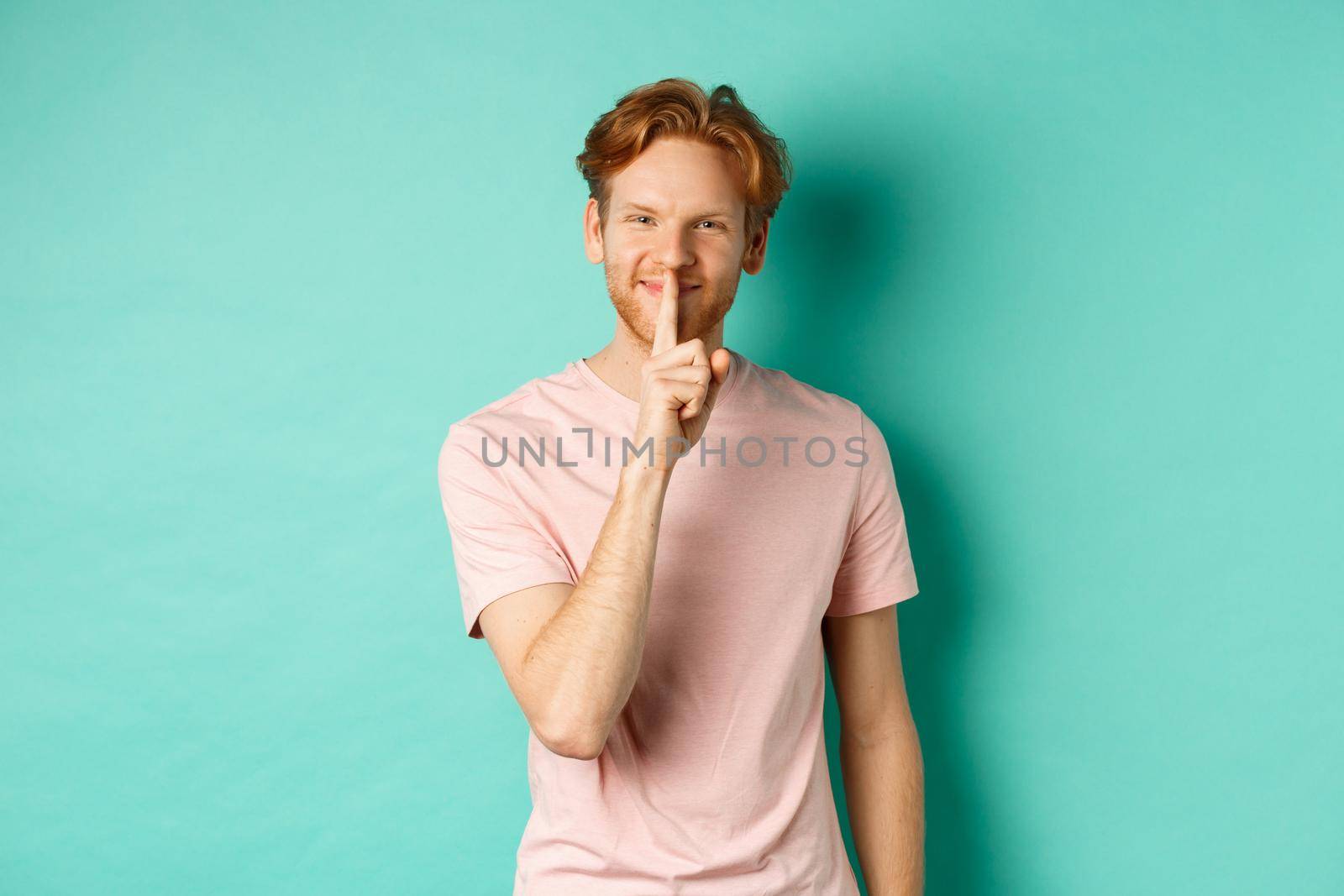 Smiling young man with red hair and beard sharing a secret, showing taboo gesture and grinning, shushing you to be quiet, standing over turquoise background by Benzoix