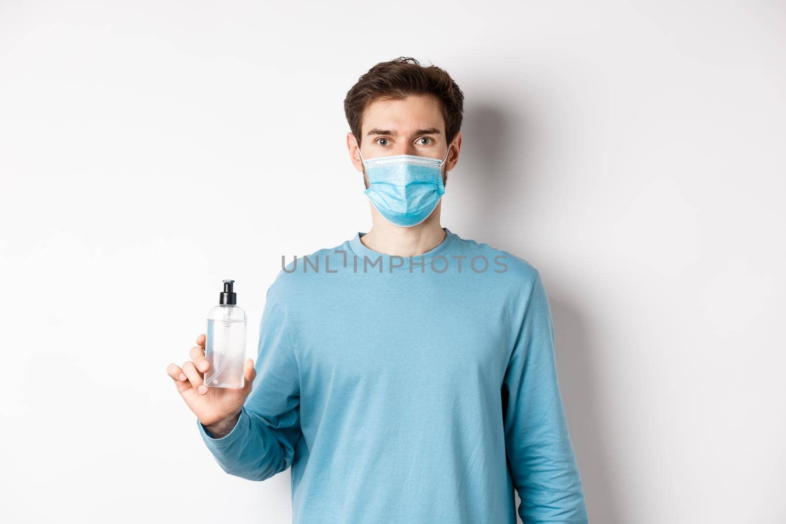 Covid-19, health and quarantine concept. Young caucasian man advice hand sanitizer, showing antiseptic and wearing medical mask, standing on white background.