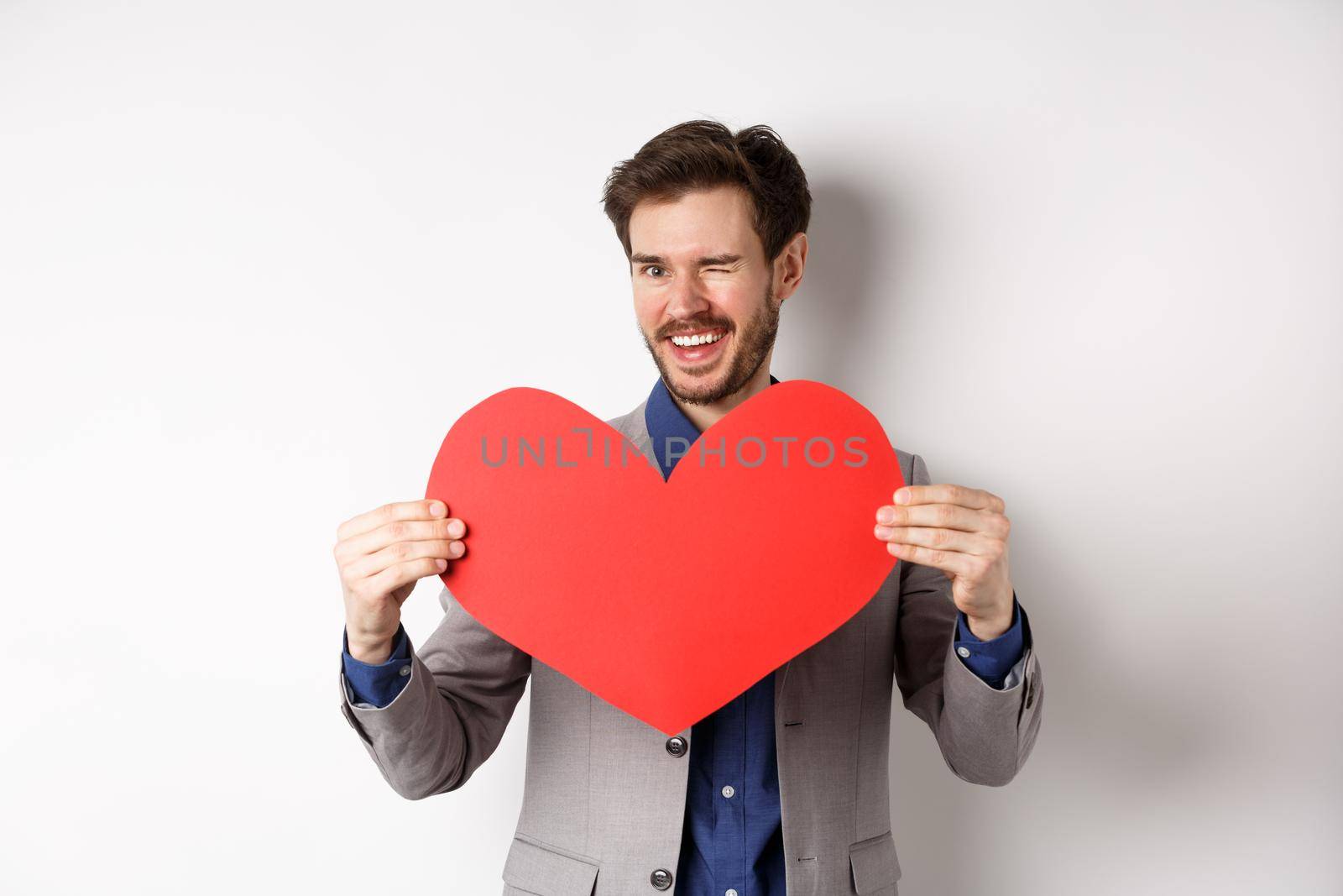 Charismatic young man winking and smiling, showing big red heart cutout for Valentines day date, say love you to lover, standing over white background by Benzoix