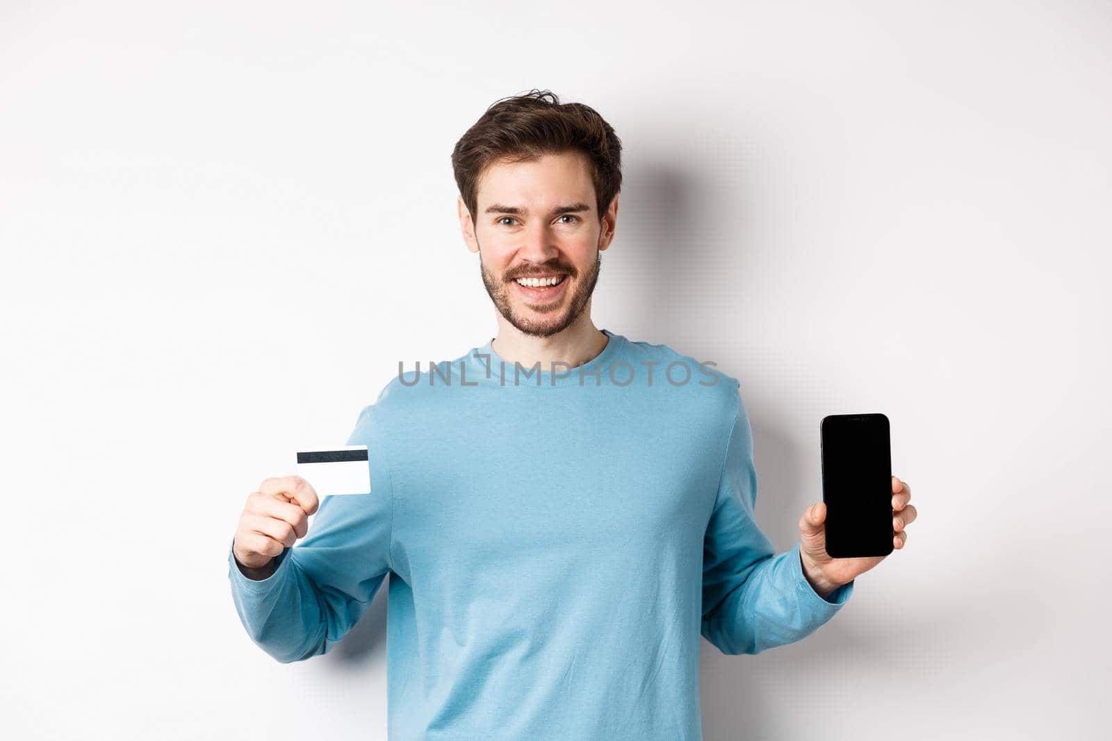 Smiling caucasian man showing plastic credit card with mobile phone screen. Guy recommending online banking app, standing on white background by Benzoix