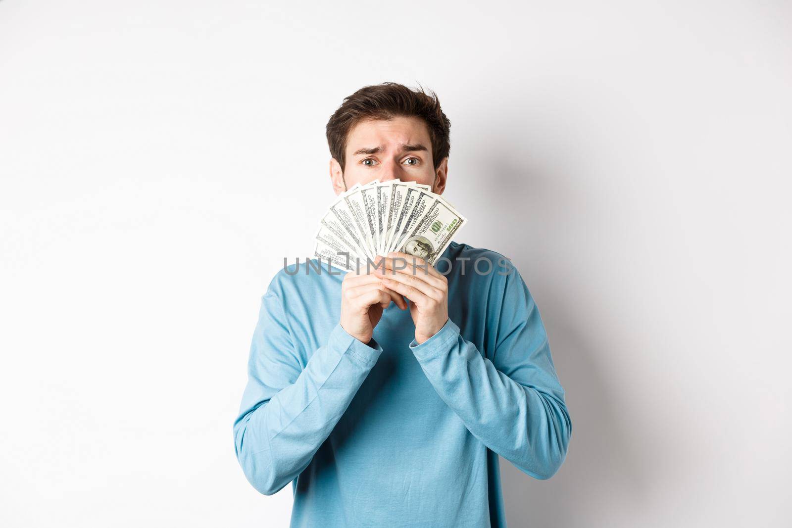 Worried young man looking with pleading face, showing money, standing in blue sweatshirt over white background.