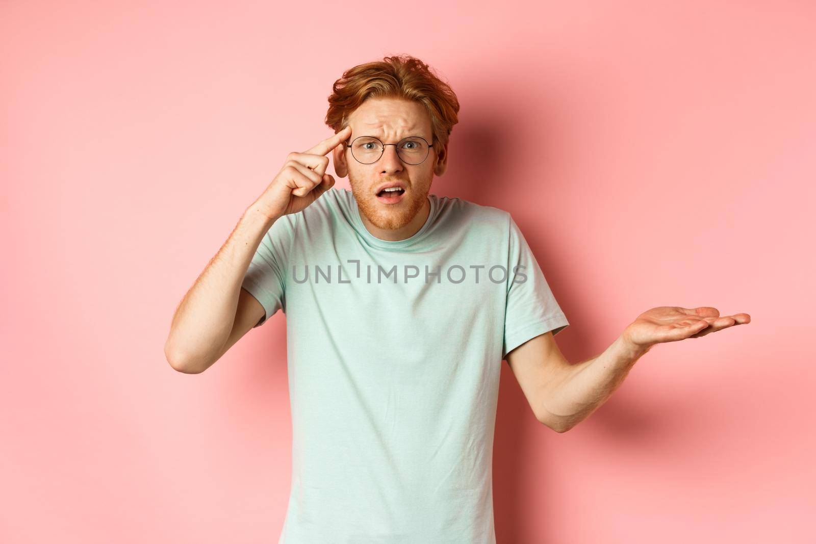 Annoyed redhead man scolding someone stupid, pointing finger at head and shrugging, standing over pink background.