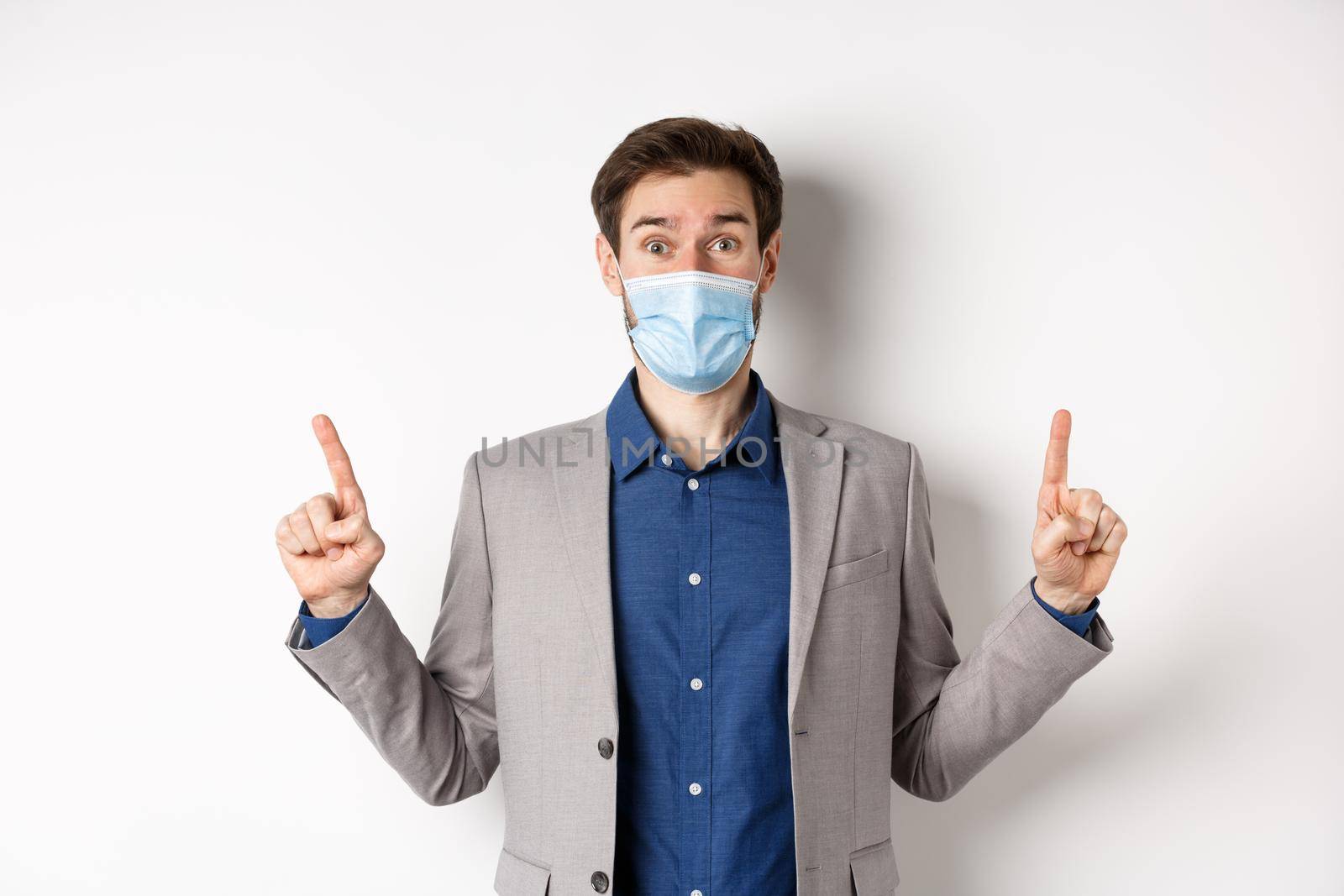 Covid-19, pandemic and business concept. Excited businessman in sterile medical mask and suit pointing fingers up, white background by Benzoix