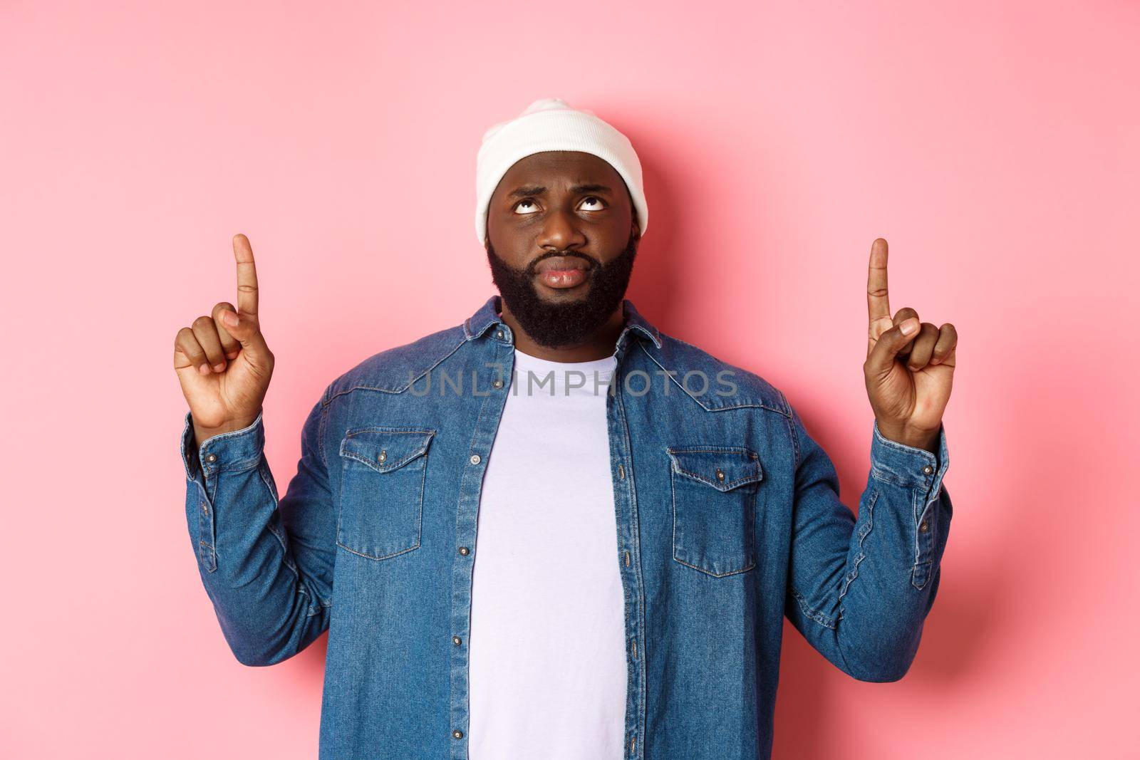 Concerned and hesitant Black man in beanie pointing fingers up, staring at top with skeptical face, standing over pink background by Benzoix