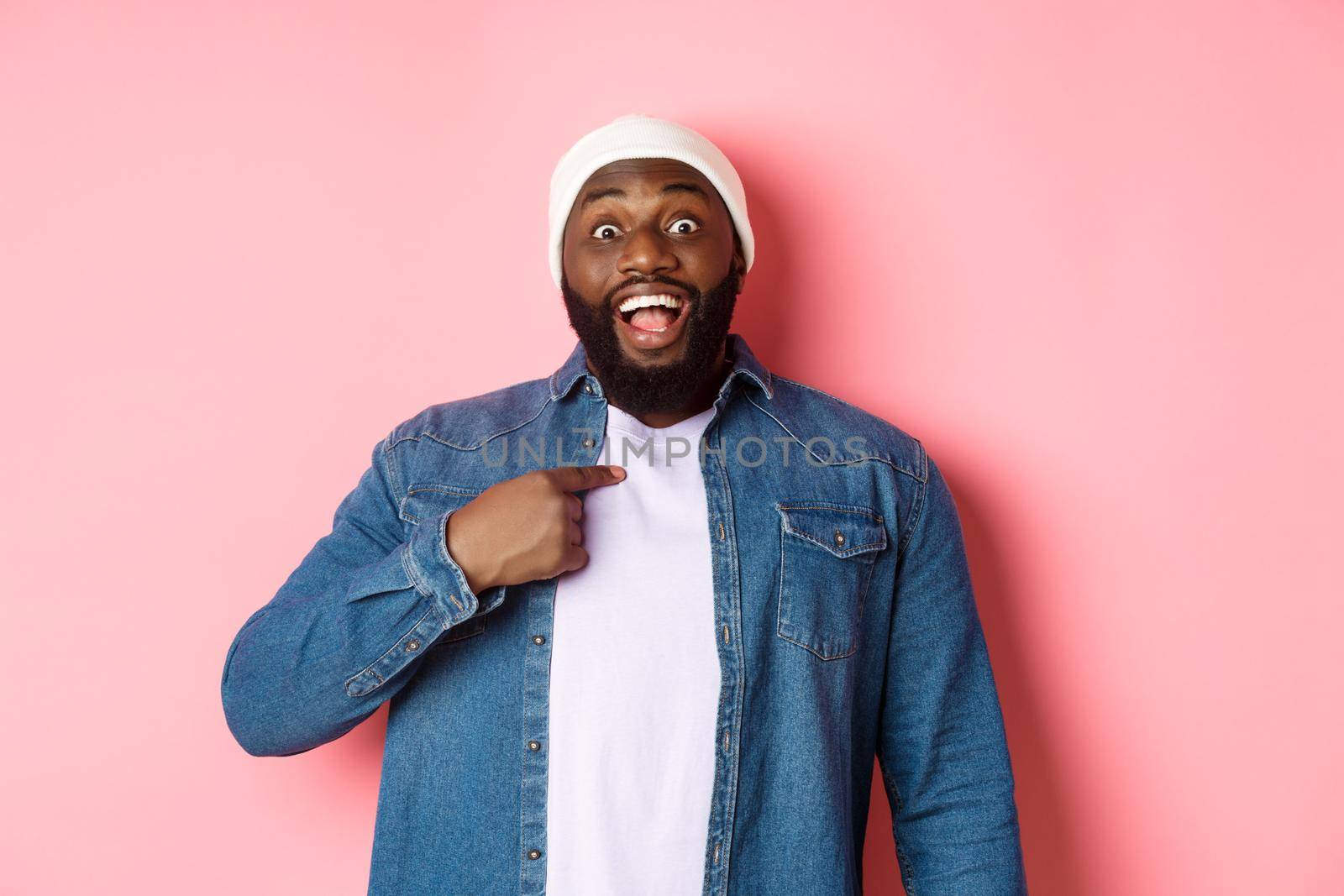 Surprised Black man in hipster beanie pointing at himself, staring at camera amazed, standing over pink background.