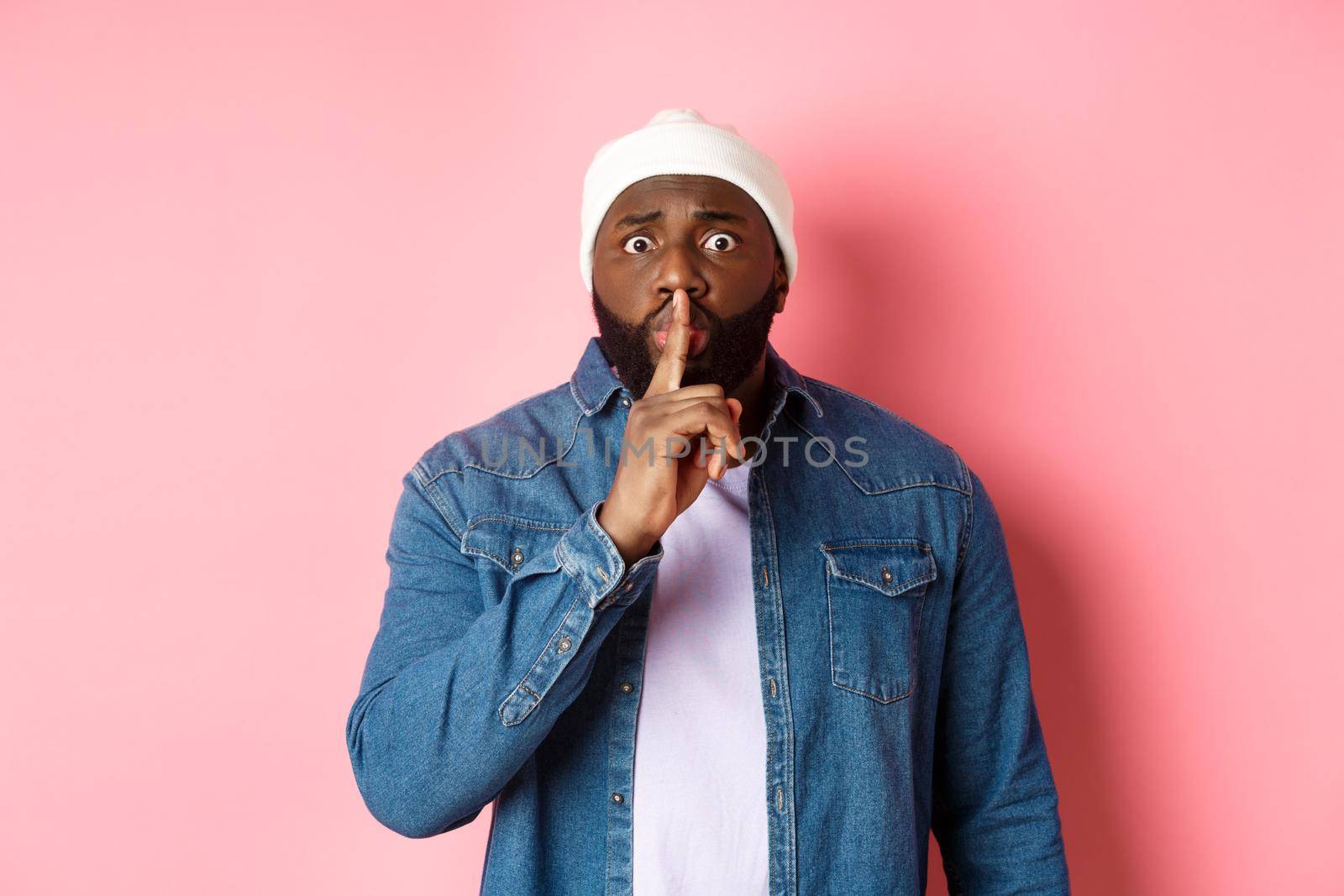 Worried Black man asking to keep quiet, sharing secret and hushing at you, holding finger pressed to lips and staring nervously at camera, pink background by Benzoix