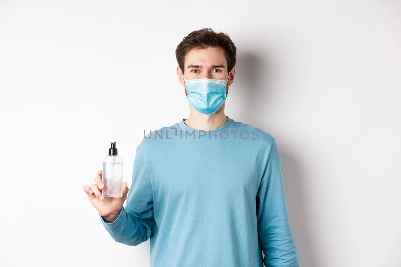 Covid-19, health and quarantine concept. Portrait of smiling man in medical mask showing hand sanitizer bottle, standing over white background by Benzoix