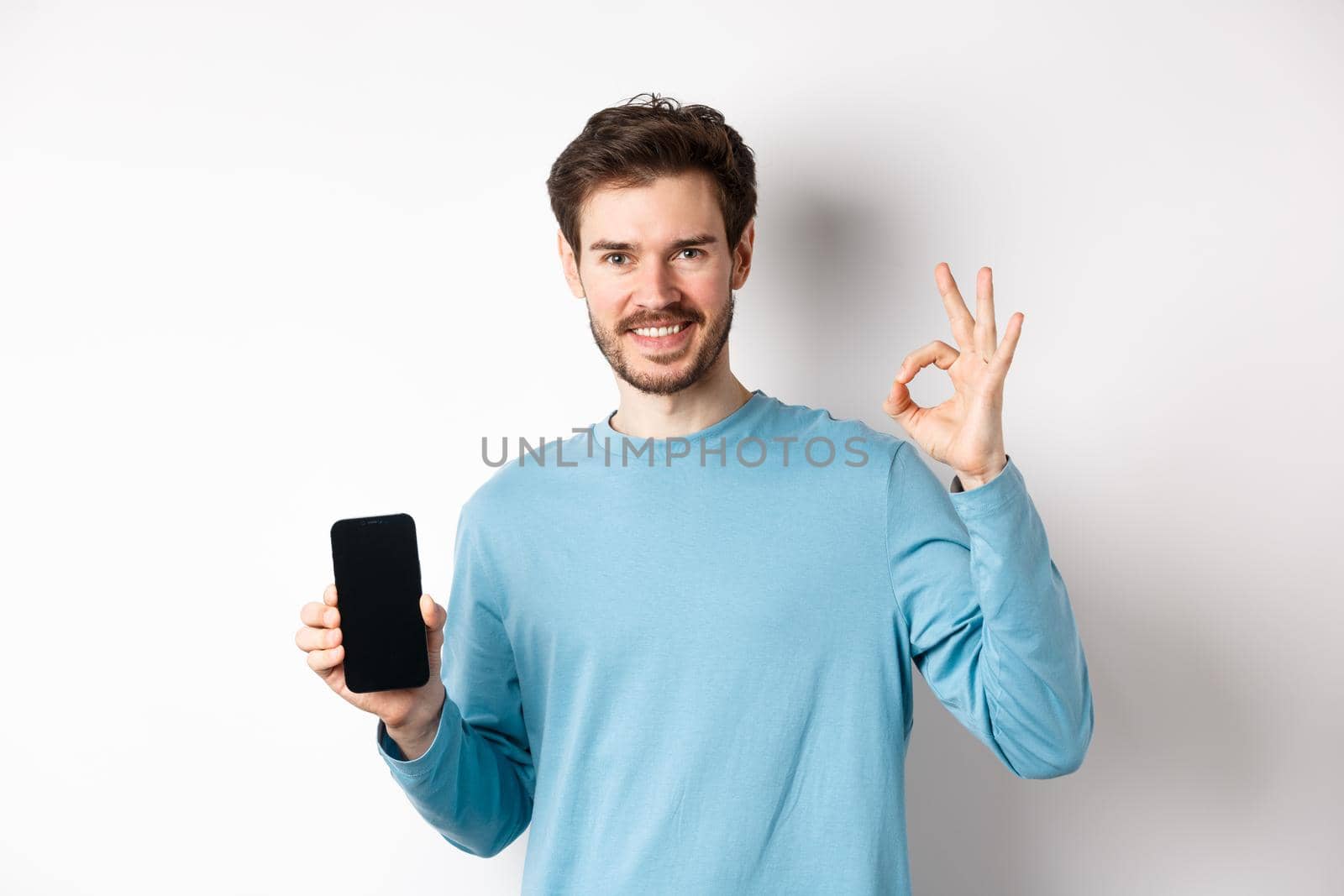 E-commerce and shopping concept. Handsome bearded guy showing empty mobile screen and okay sign, praising good app, standing over white background.