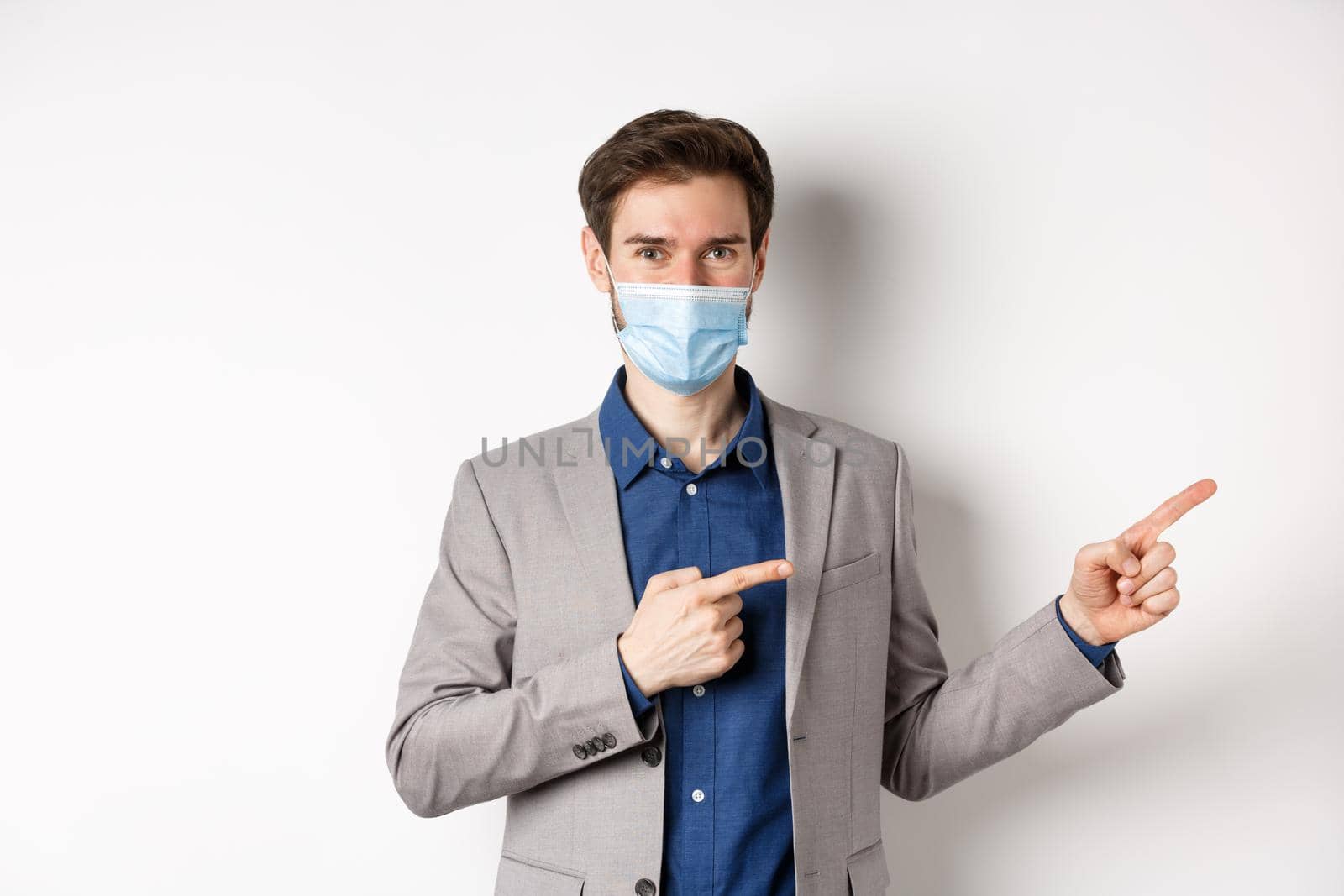 Covid-19, pandemic and business concept. Confident male manager in suit and medical mask showing way, pointing fingers right at advertisement, white background.