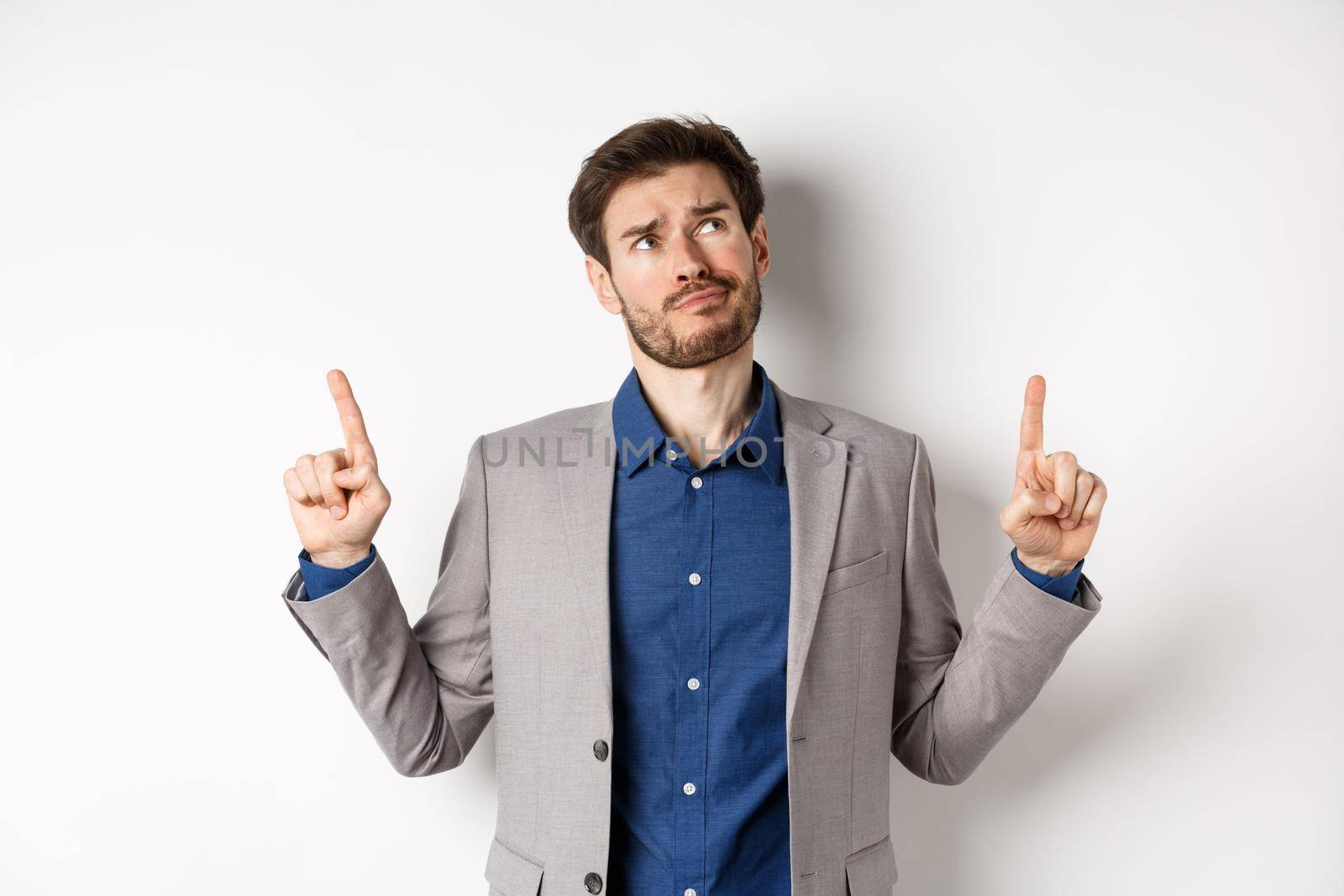 Skeptical and hesitant young businessman in suit looking up with disappointed smirk, feel unsure about deal, standing dissatisfied on white background.