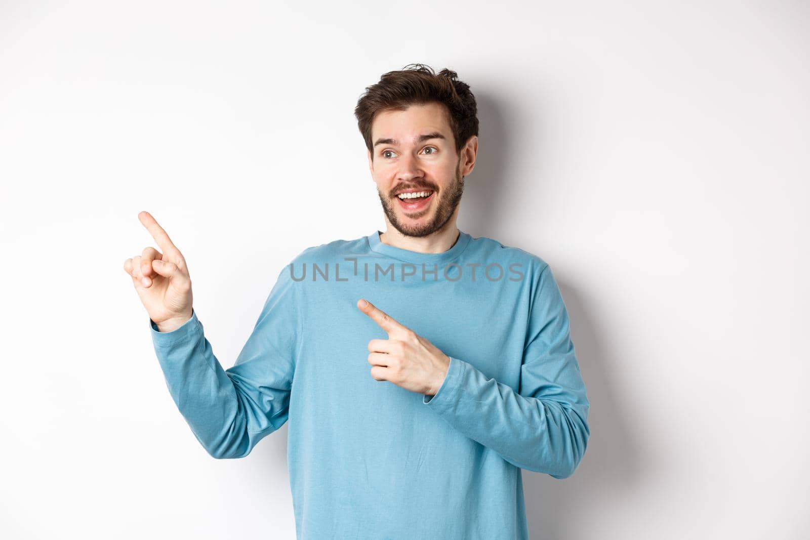 Excited bearded man checking out special offer, pointing and looking at advertisement with happy face, standing over white background by Benzoix