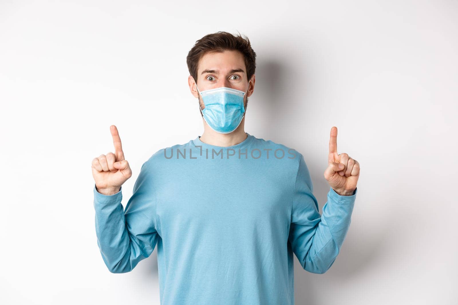 Coronavirus, health and quarantine concept. Excited smiling guy checking out special deal, pointing fingers up, stare amazed at camera, wear medical mask on white background.