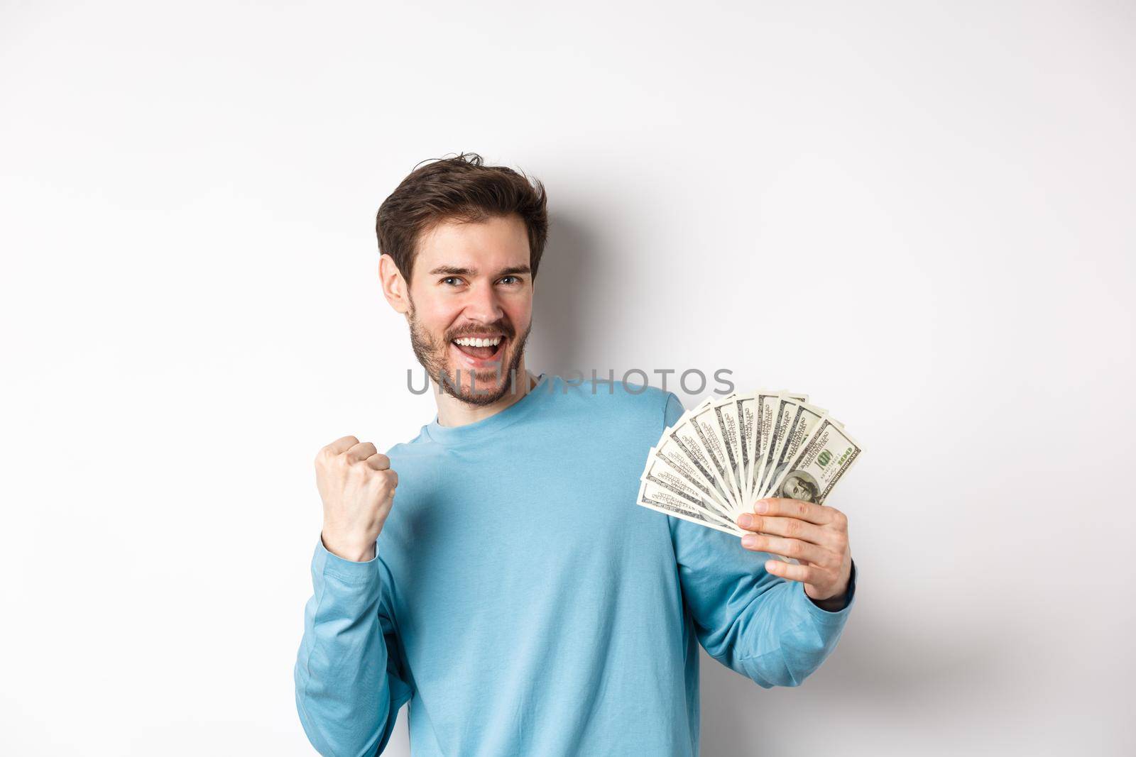 Happy man dancing with money, showing dollars and saying yes with satisfied smile, making fist pump gesture, standing over white background by Benzoix