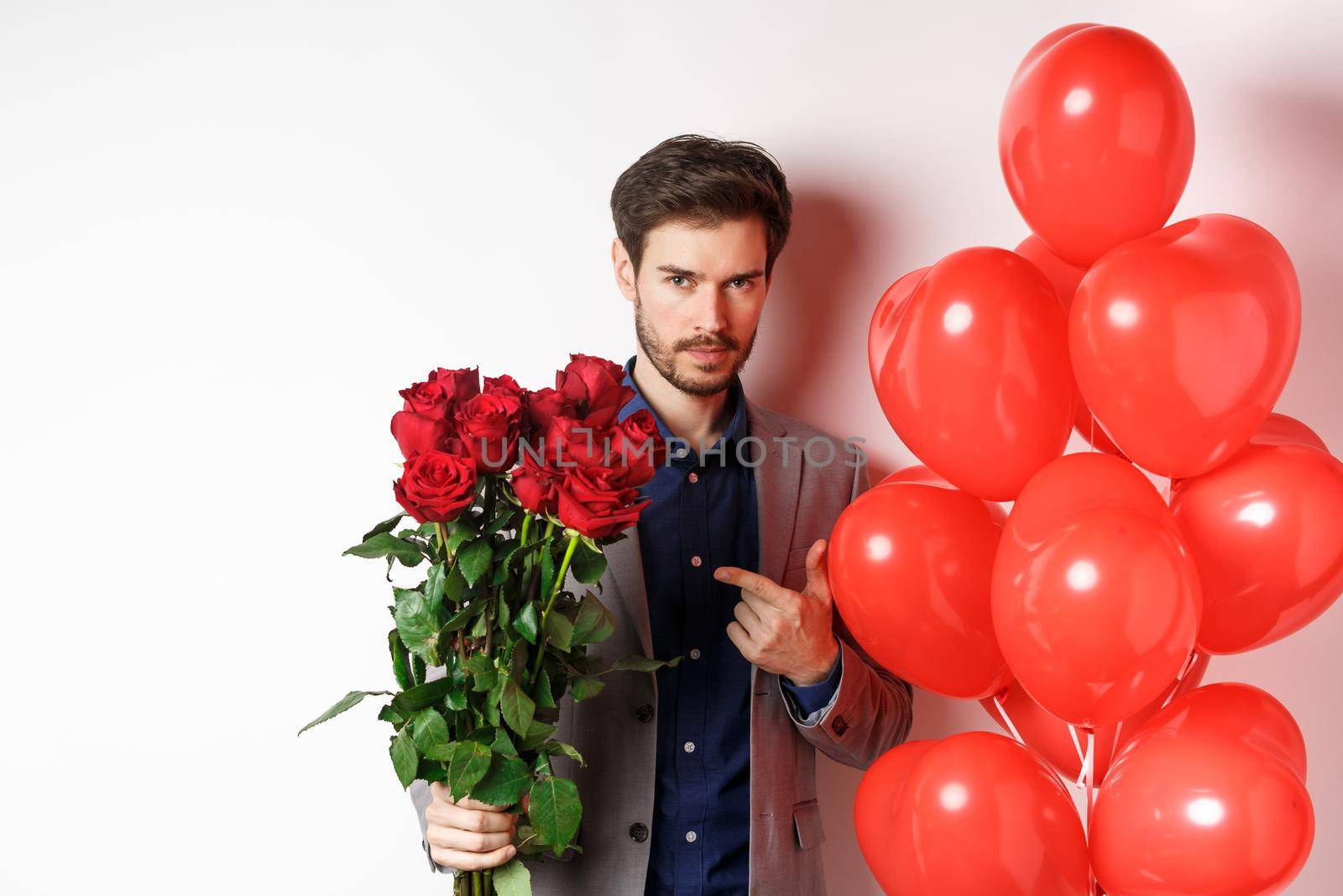 Handsome man in suit going on romantic Valentines day date, pointing at bouquet of roses, standing near heart balloon over white background by Benzoix