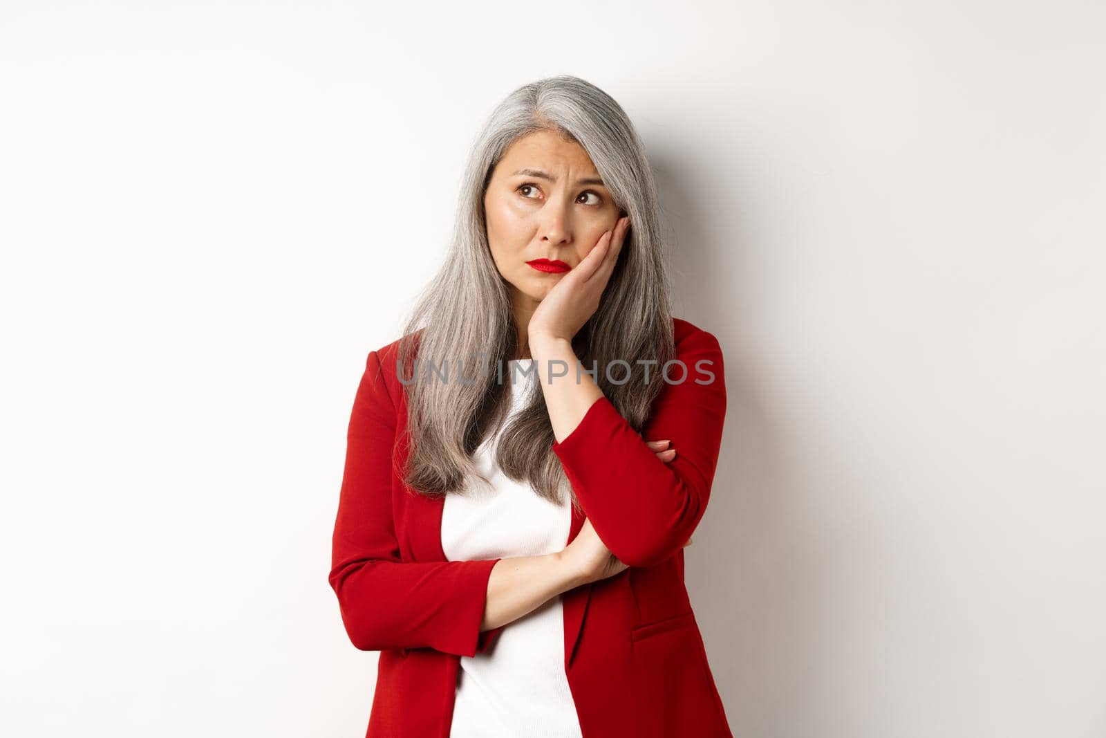 Troubled and sad asian female entrepreneur looking at upper left corner, thinking with upset face, standing over white background by Benzoix