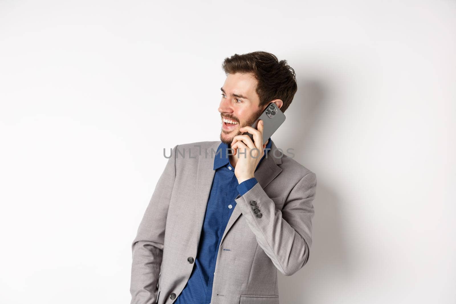 Happy businessman laughing and smiling, talking on mobile phone, holding hand in pocket and looking aside at empty space with cheerful face, white background.