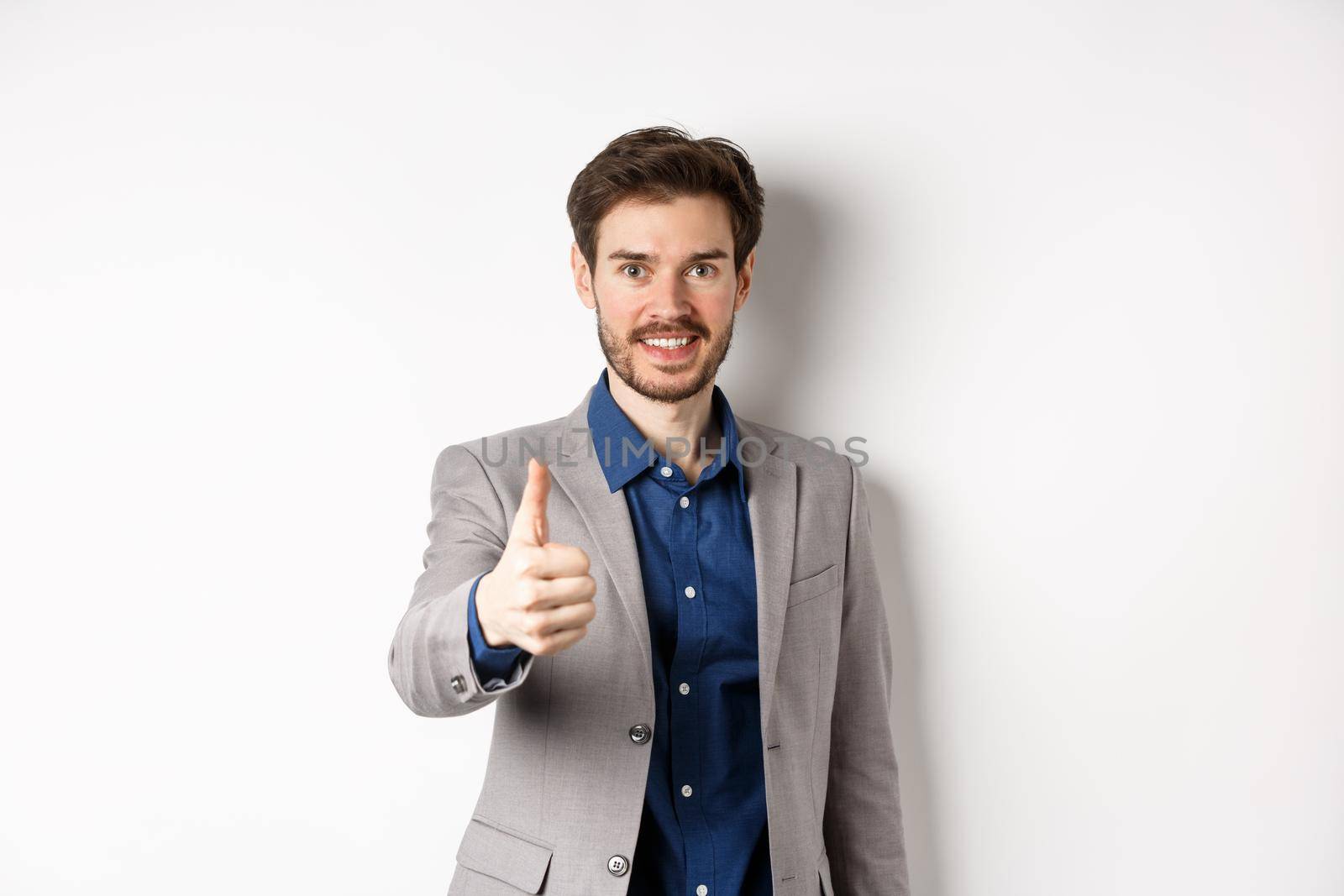 Image of handsome businessman in suit showing thumb up and smiling, standing on white background by Benzoix