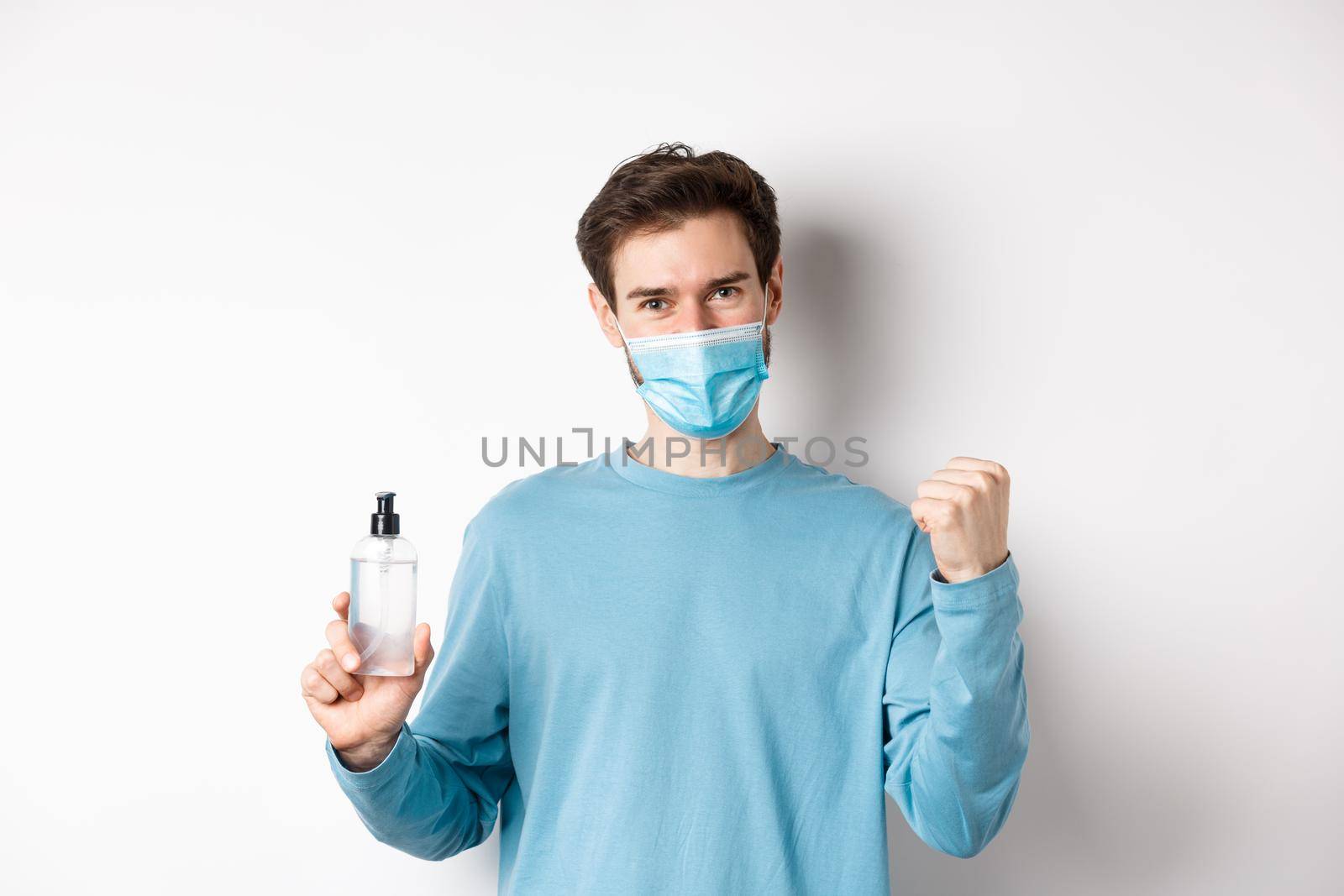 Covid-19, health and quarantine concept. Cheerful man in face mask celebrating, showing fist pump and bottle with hand sanitizer, fighting germs, white background.