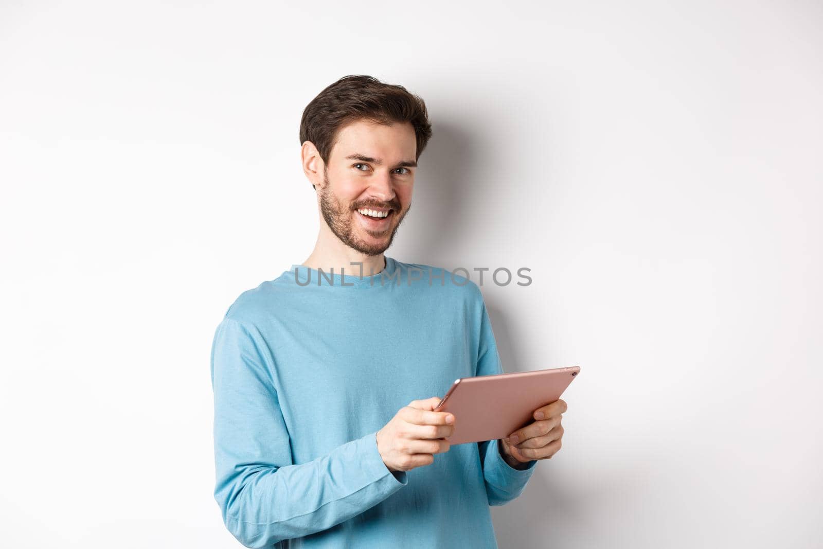 E-commerce and technology concept. Young caucasian man smiling at camera, holding digital tablet, standing over white background by Benzoix
