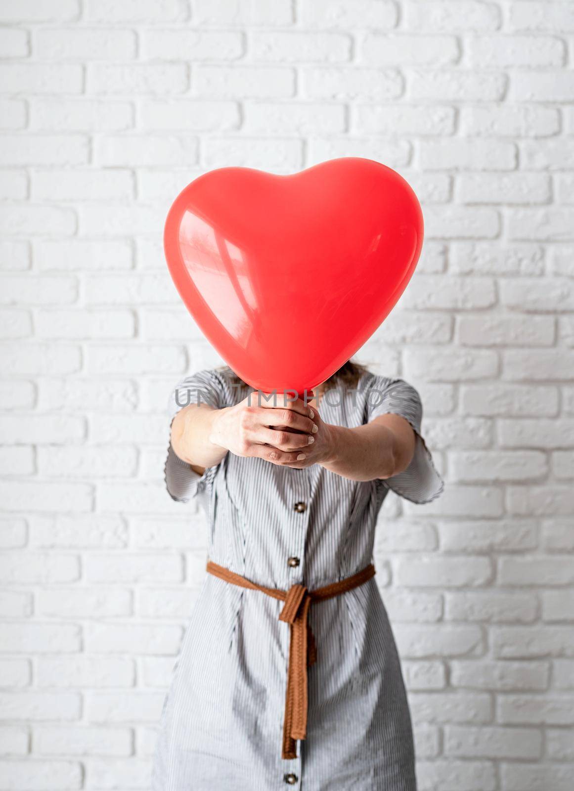 Valentines Day. Young woman holding read heart shaped balloon on white brick wall background