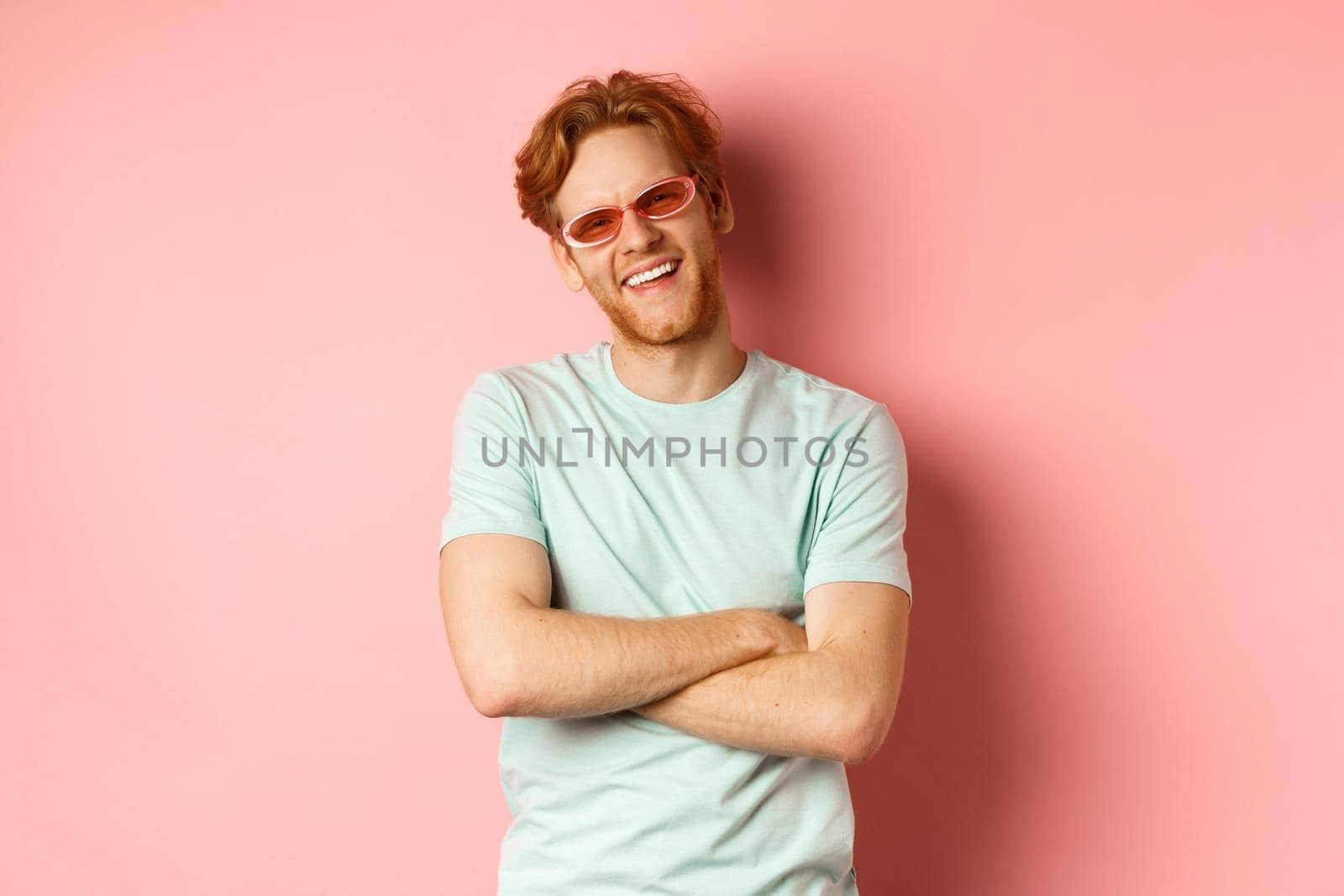 Tourism and vacation concept. Friendly young man with red hair, wearing sunglasses and t-shirt, cross arms on chest and smiling joyfully at camera, pink background.