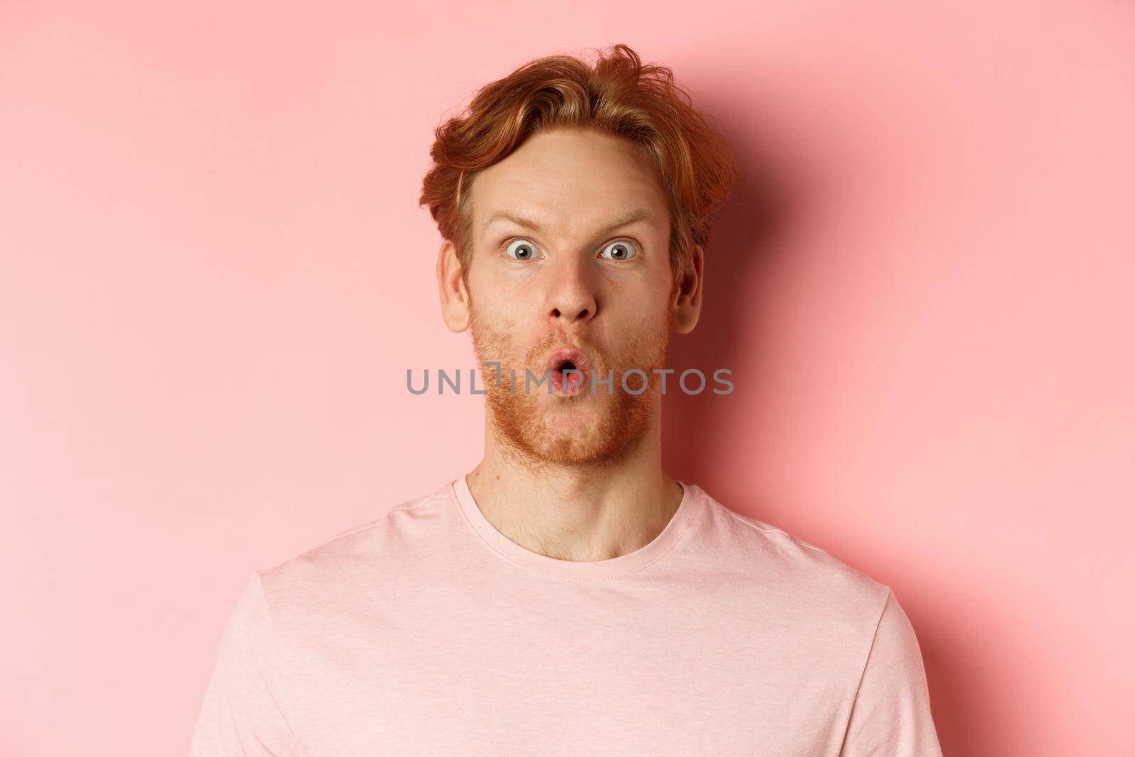 Headshot of young redhead man with beard, saying wow and staring at camera amazed, checking out promo deal, standing over pink background by Benzoix