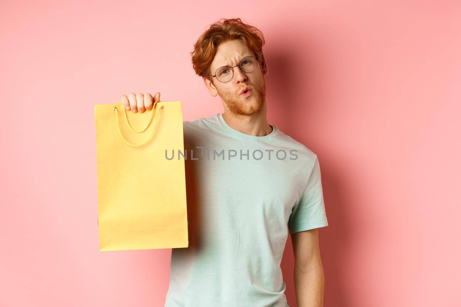 Handsome young man in glasses looking sassy at camera, showing yellow shopping bag, telling about discounts in shop, standing over pink background.