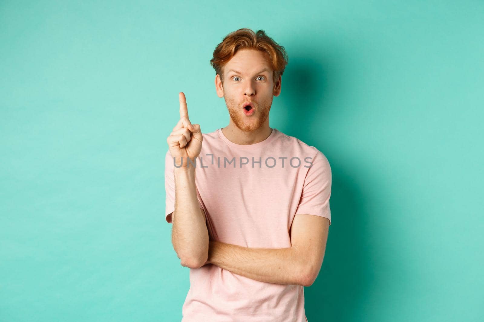 Pensive redhead man in t-shirt raising index finger, gasping as pitching at idea, saying suggestion, standing over turquoise background. Copy space