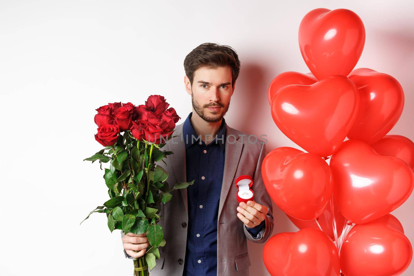Handsome young man prepare to make proposal, holding engagement ring with bouquet of red roses, making surprise on Valentines day, standing near heart balloons, white background.