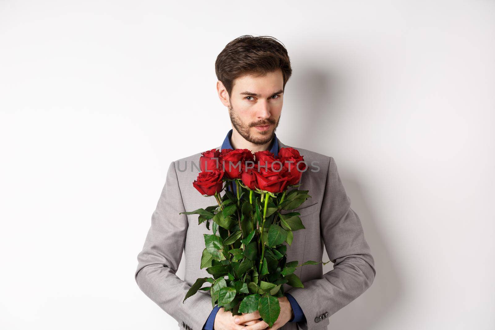 Handsome macho man going on date in suit, holding bouquet of red roses and smiling at camera, making valentines day surprise gift, white background by Benzoix