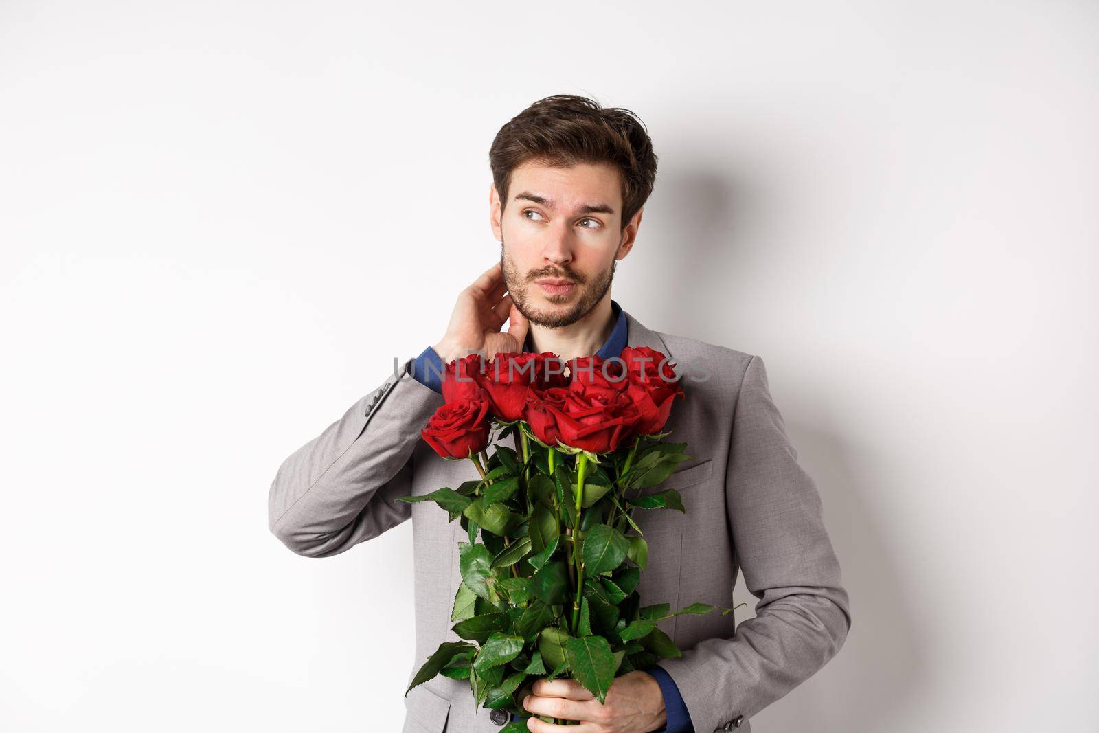 Pensive young man in suit holding bouquet of flowers, waiting for date on Valentines day, standing over white background.