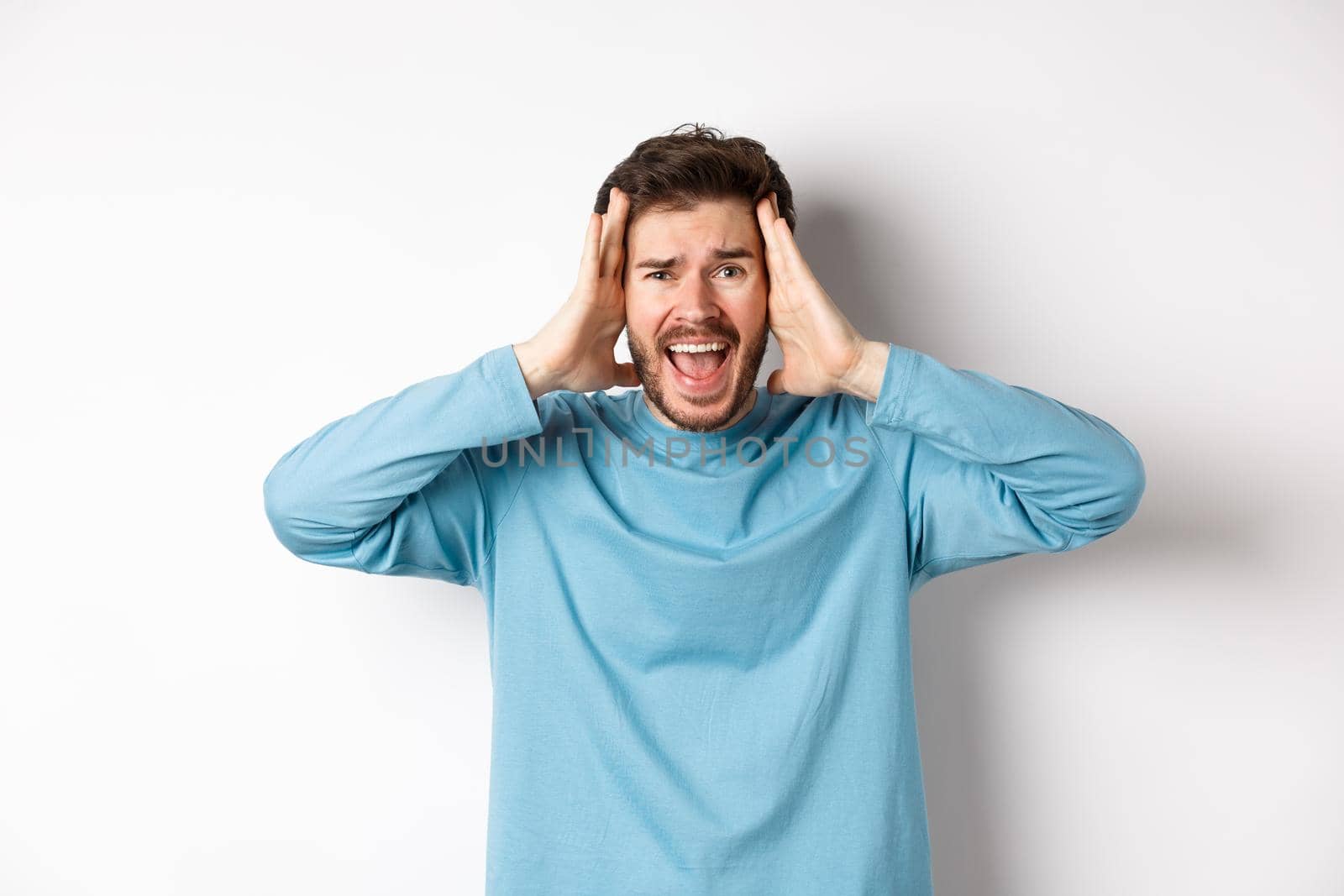 Image of young caucasian man screaming in panic, looking frustrated and shouting at camera, standing on white background.