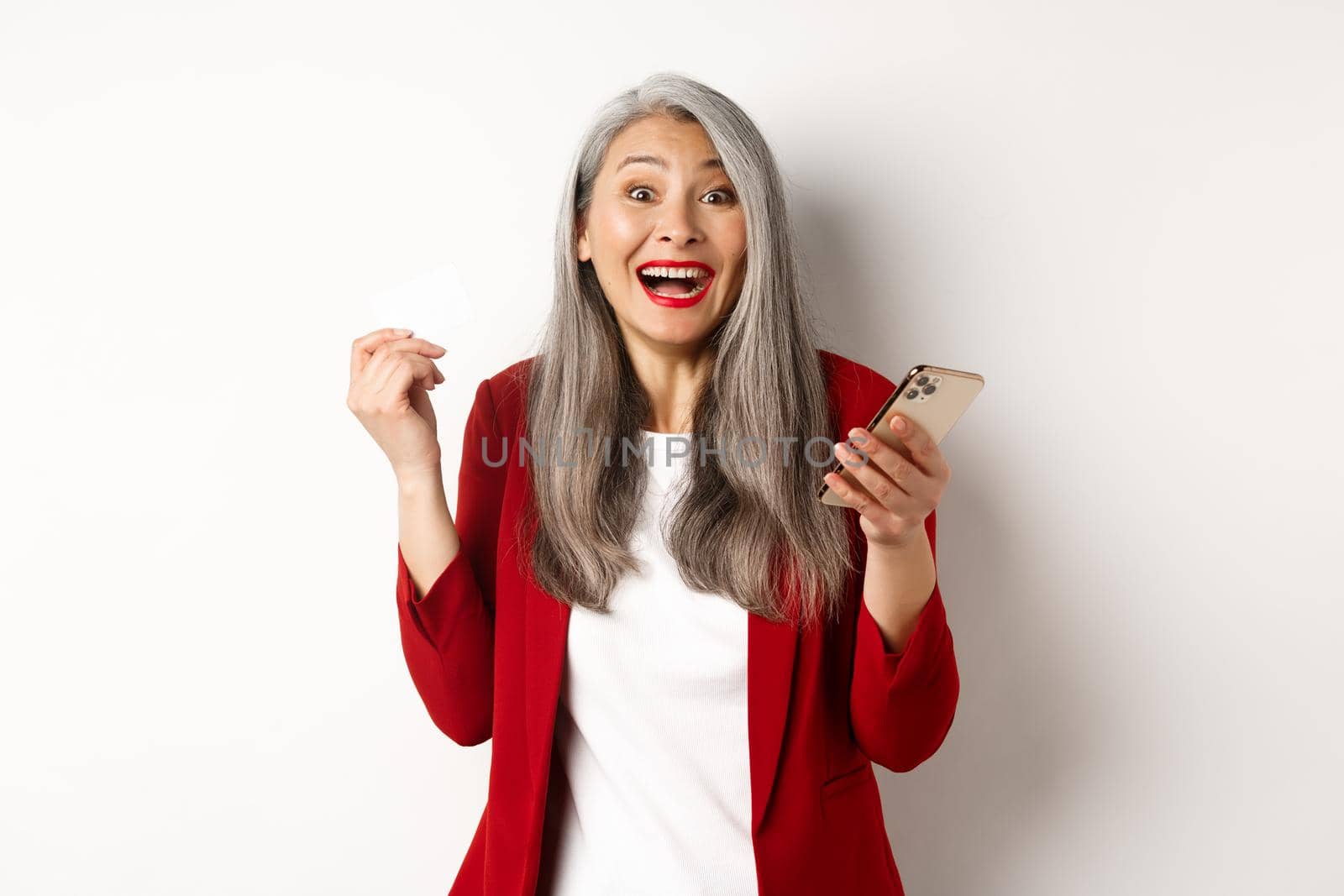 Excited asian businesswoman in red blazer, holding plastic credit card and smartphone, smiling happy at camera, standing over white background by Benzoix