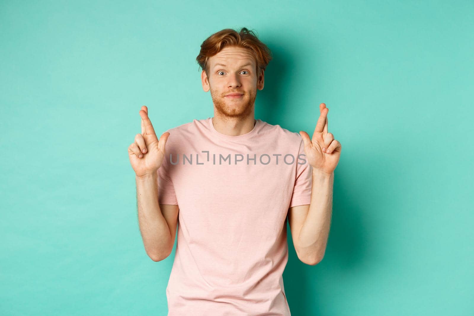 Smiling hopeful man with red hair making a wish, cross fingers for good luck and expecting something good, standing over turquoise background.