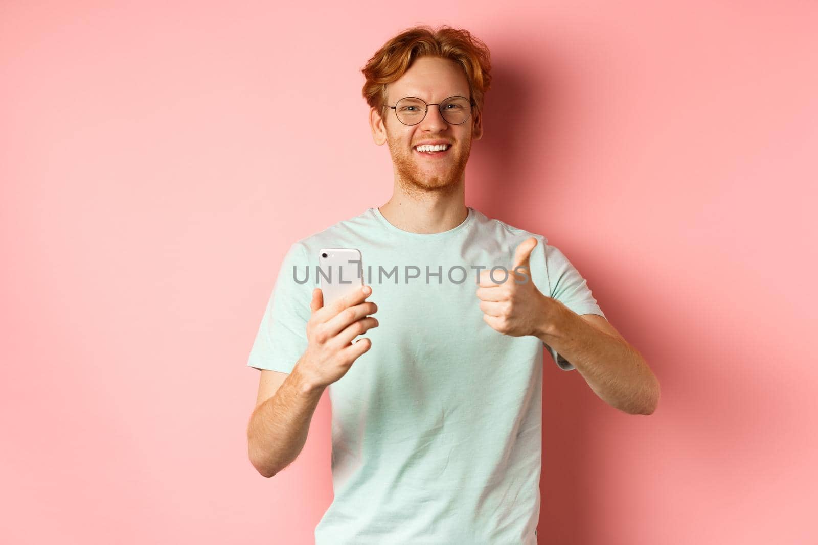 Happy young man with red messy hair, showing thumbs-up while using mobile phone, smiling and praising application, standing over pink background.
