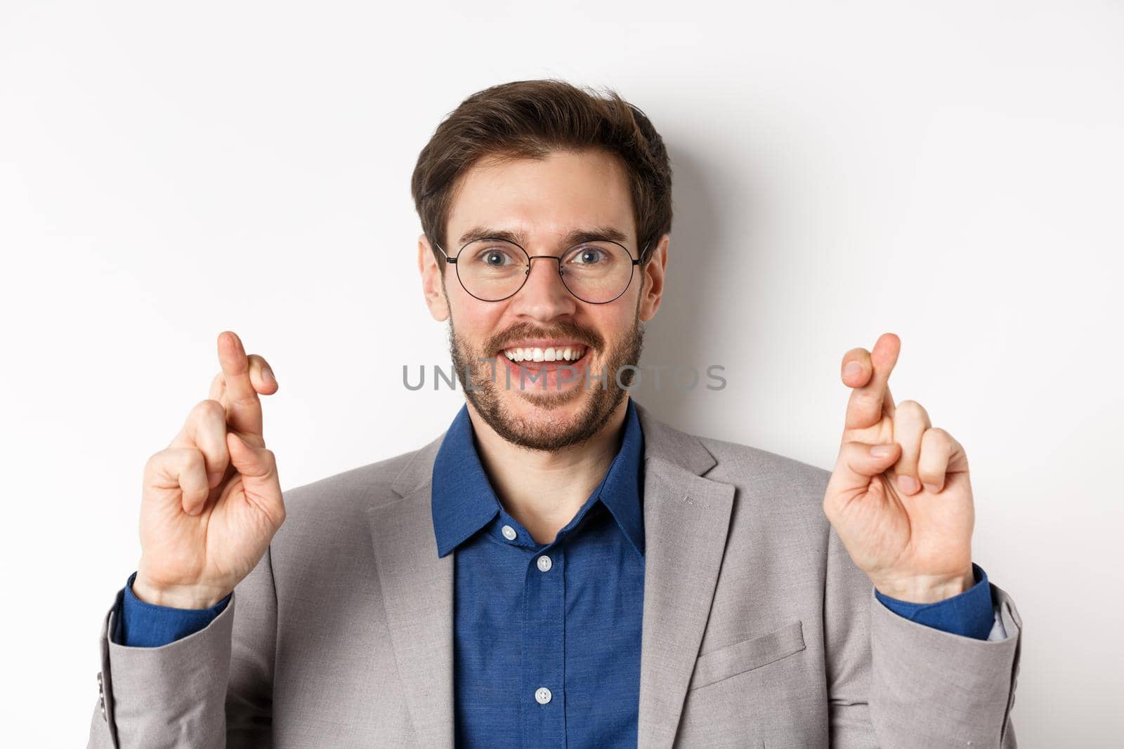 Close-up of hopeful male entrepreneur in glasses and suit cross fingers for good luck, smiling and making wish, praying for good results, white background.