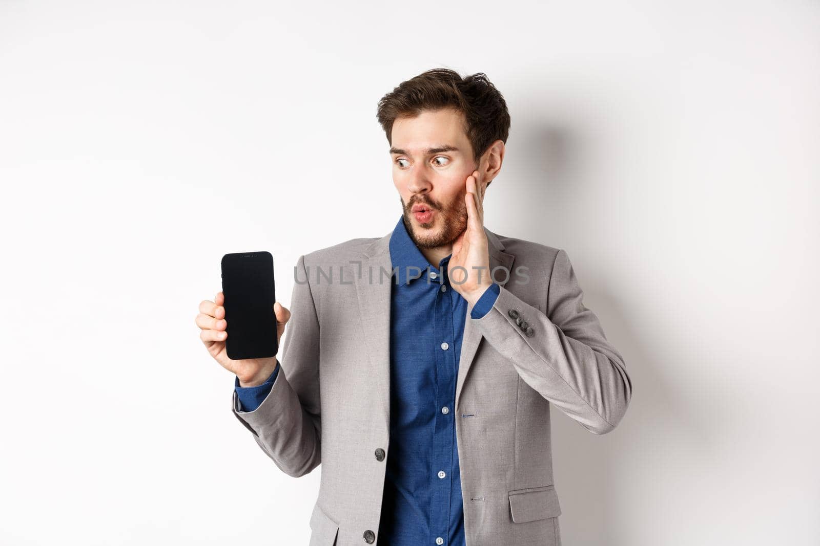 E-commerce and online shopping concept. Amazed male manager in suit looking at mobile phone screen with excited face, standing on white background.