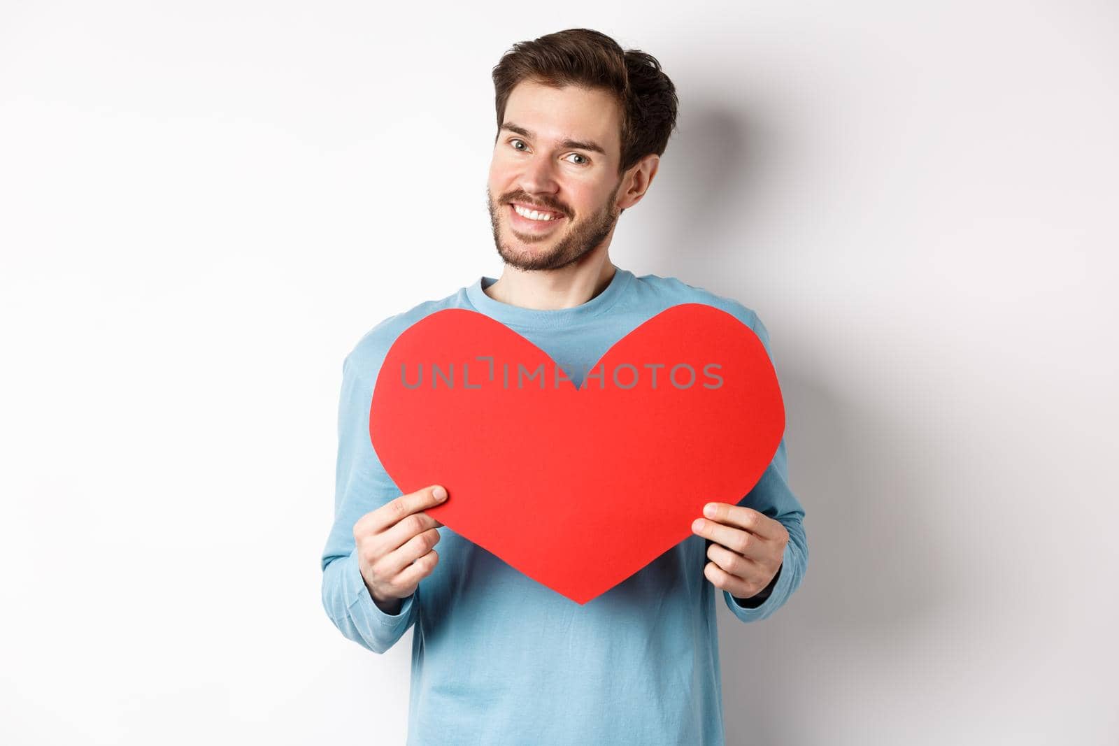 Handsome european man in sweater saying I love you, boyfriend standing with valentines day red heart, posing over white background.