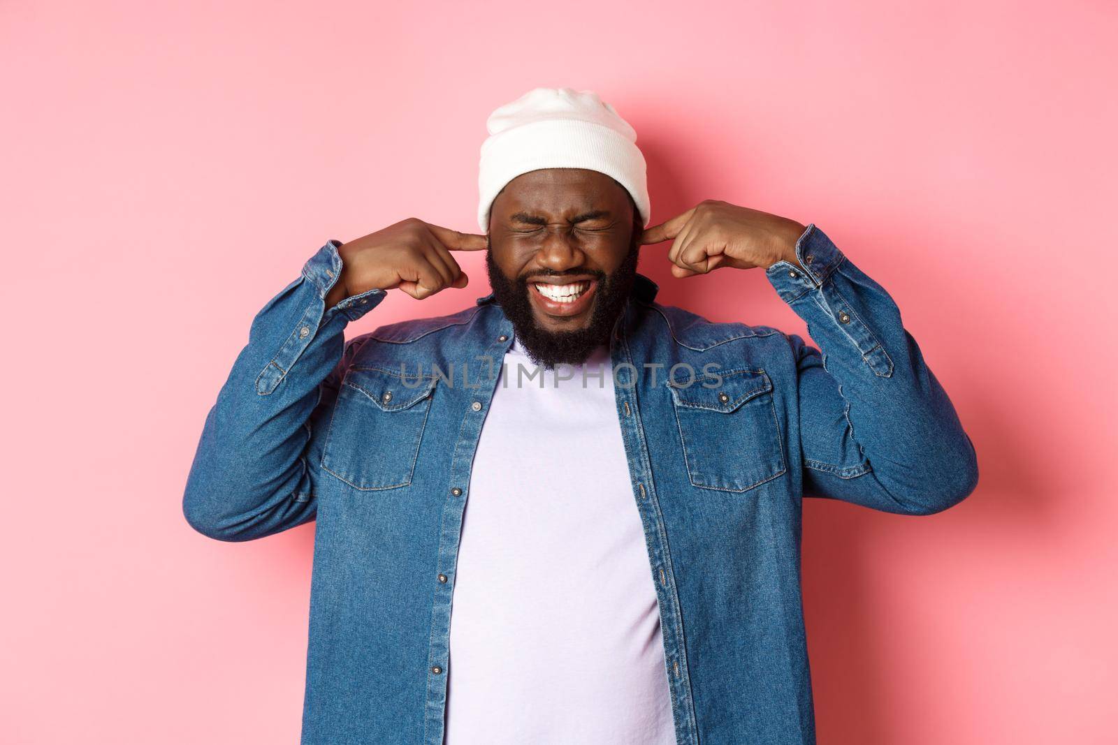 Displeased african-american man shut ears and grimacing from loud awful noise, disturb by annoying sound, standing over pink background.