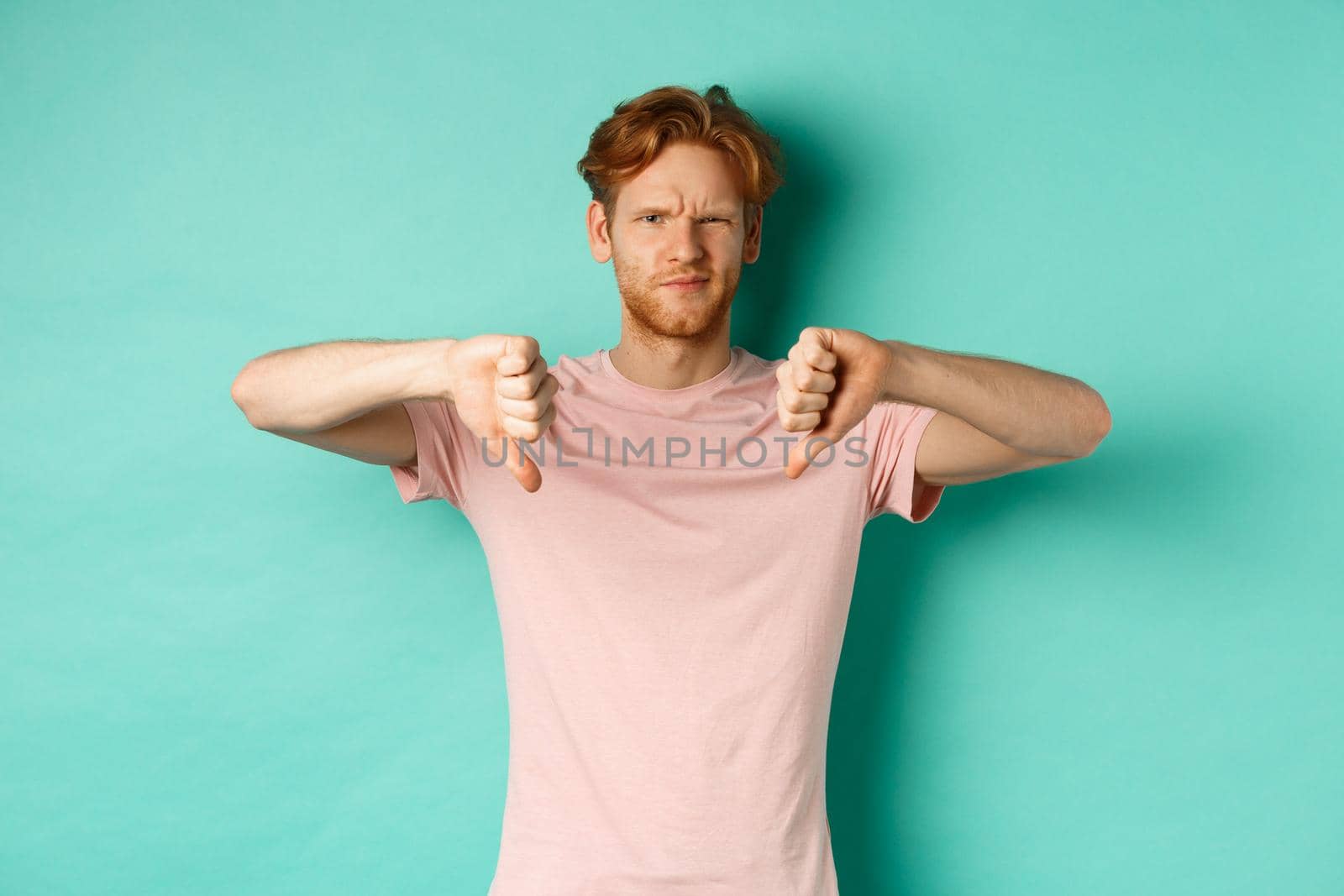 Disappointed guy with red hair showing thumbs-down, frowning and looking skeptical, epxress dislike and disapproval, standing over turquoise background.