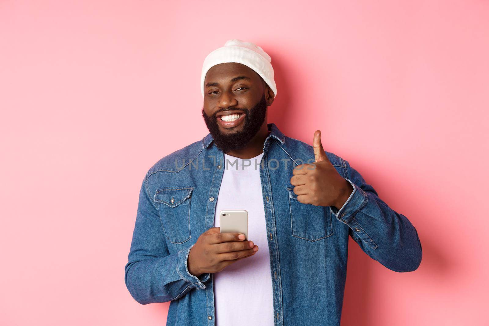 Technology and online shopping concept. Happy african-american guy showing thumb-up in approval while using mobile phone, standing satisfied over pink background.