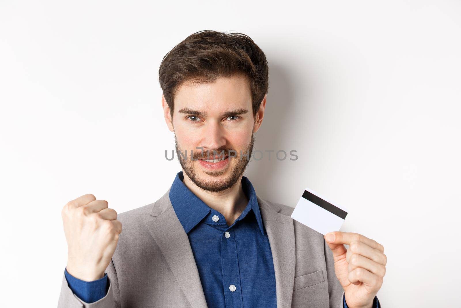 Shopping. Cheerful handsome businessman showing plastic credit card and fist pump, saying yes with rejoice, making money, standing on white background.