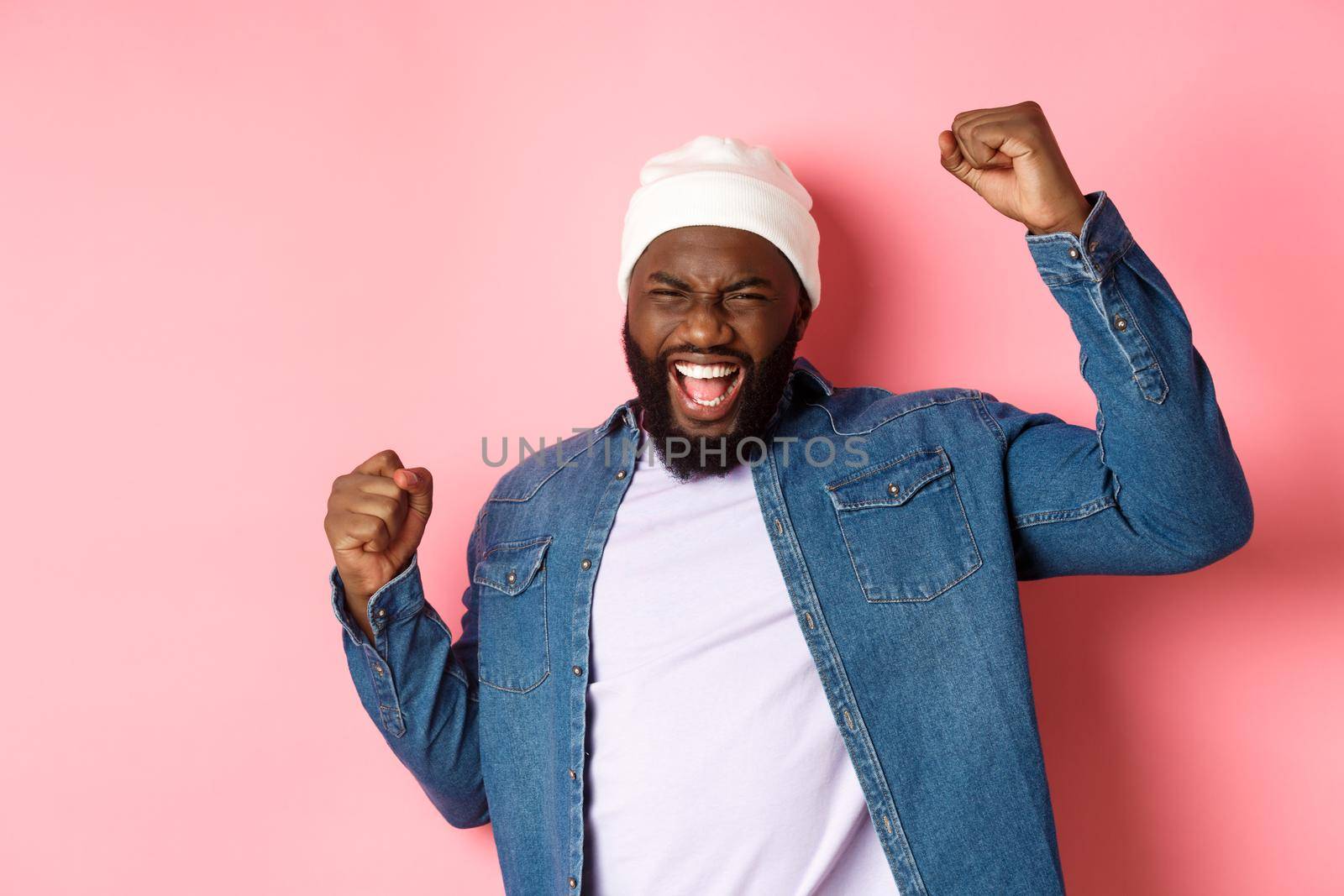 Lucky african-american guy winning prize, raising hands up in triumph gesture, scream of joy yes, celebrating victory, pink background by Benzoix