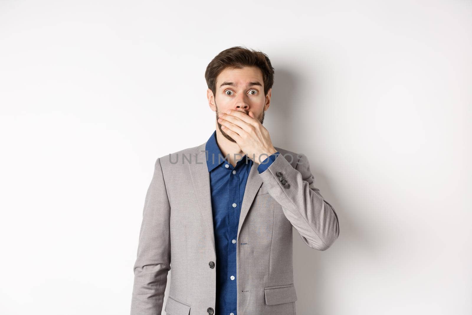Shocked boyfriend in suit gasping, covering mouth with hand and pop eyes at camera startled, standing on white background by Benzoix
