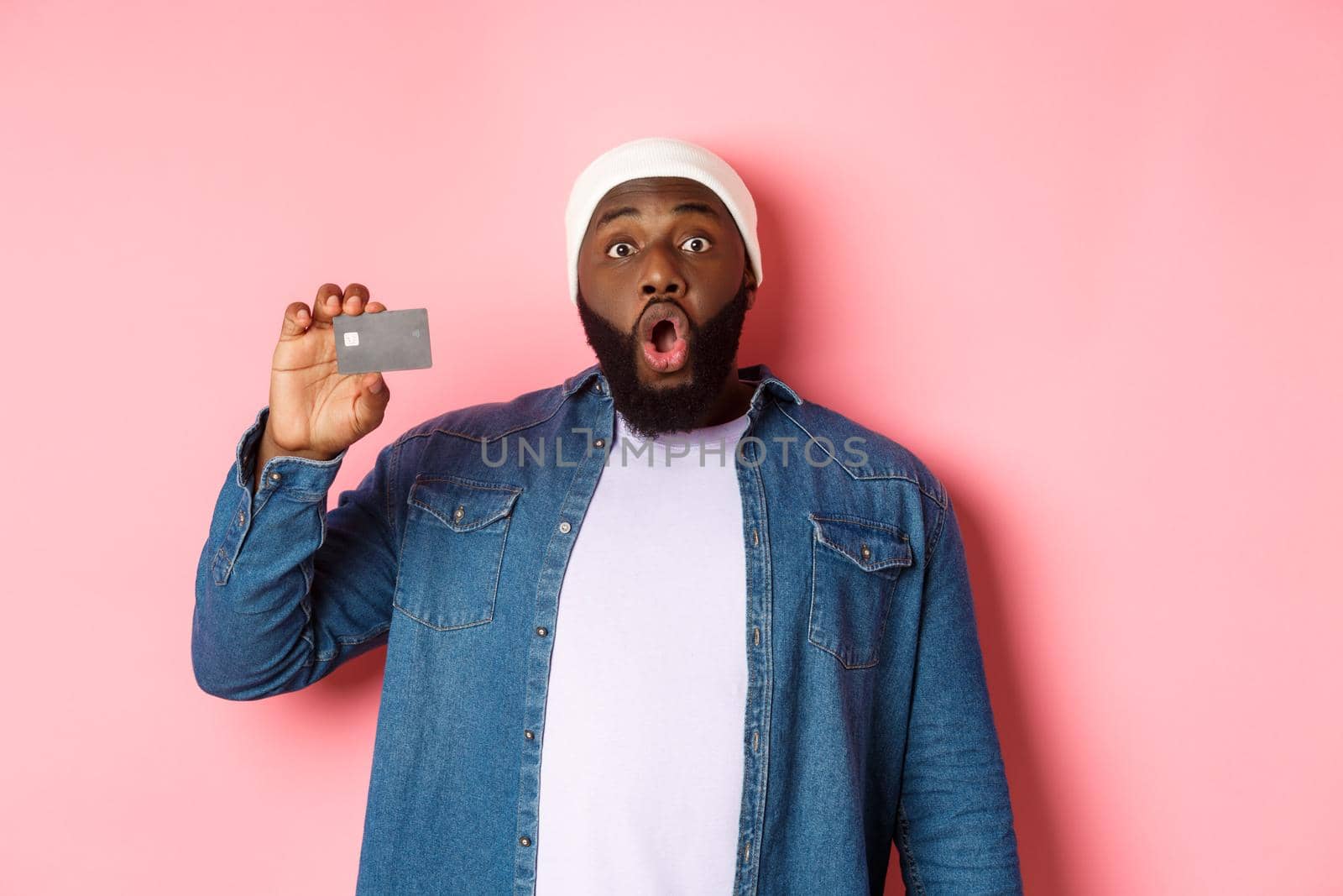 Shopping concept. Amazed Black guy showing credit card, staring at camera impressed, recommending bank, standing over pink background.